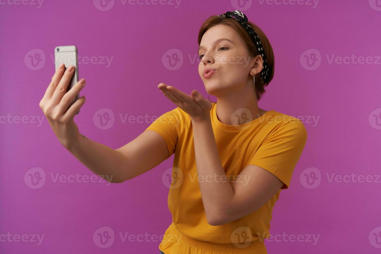 ritratto di attraente giovane Marrone capelli donna con naturale trucco indossare giallo camicia e nero bandana emozione flirtare amorevole e Spedire aria bacio rendere tiro su Telefono in posa contro viola sfondo foto