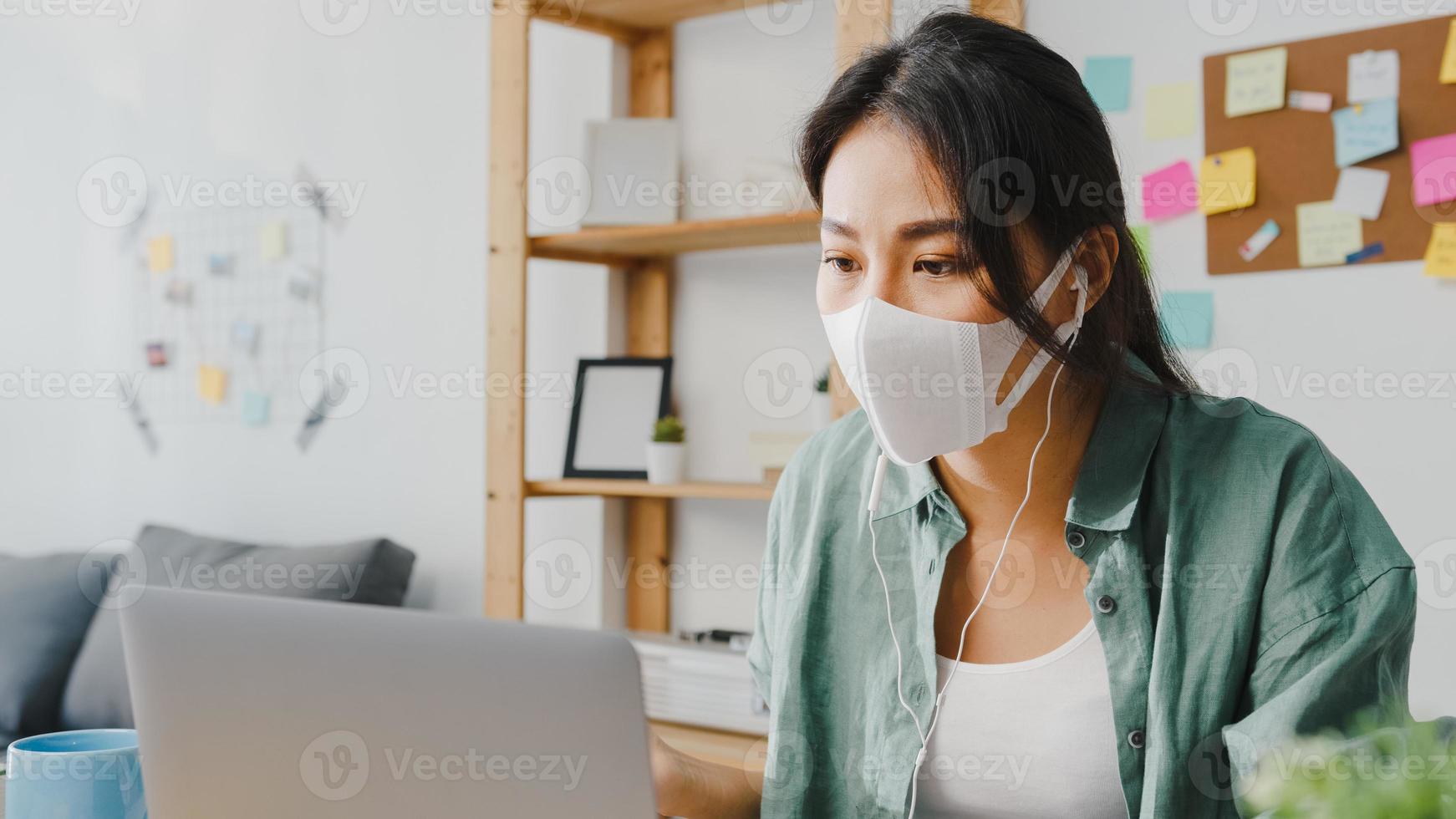 donna d'affari asiatica che indossa una maschera medica utilizzando il laptop parla con i colleghi del piano in videochiamata mentre lavora da casa in soggiorno. distanziamento sociale, quarantena per la prevenzione del virus corona. foto