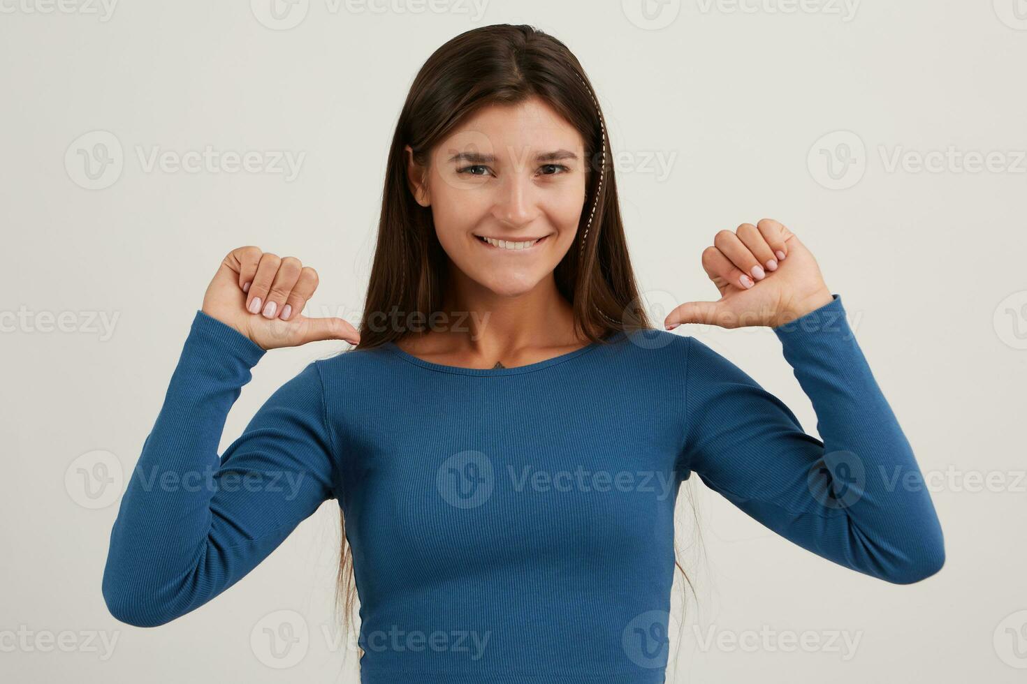 giovane signora, bella donna con buio lungo capelli. indossare blu Maglione. emozione concetto. puntamento pollici a se stessa. pungente labbro. Guardando a il telecamera isolato al di sopra di bianca sfondo foto