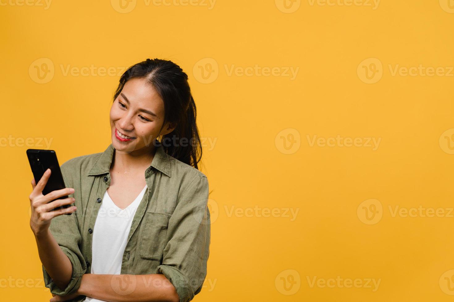 giovane donna asiatica che usa il telefono con un'espressione positiva, sorride ampiamente, vestita con abiti casual sentendosi felici e in piedi isolato su sfondo giallo. felice adorabile donna felice esulta successo. foto