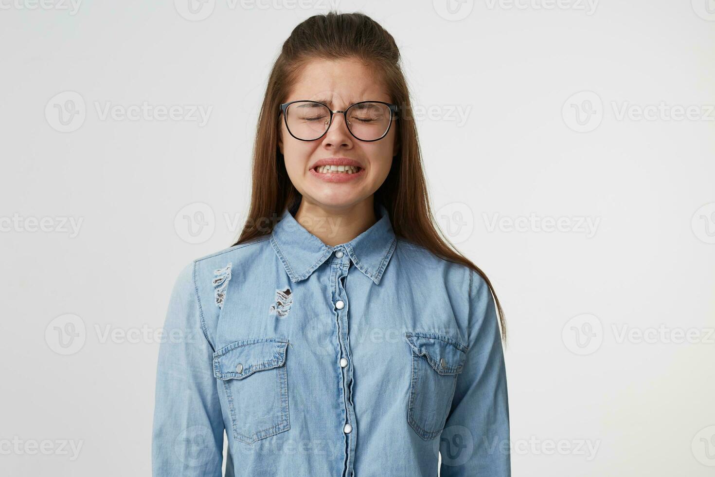 molto irritato giovane donna nel bicchieri in piedi con occhi chiuso pianto stringendo sua denti apertura sua bocca si sente indifeso indossare un' denim camicia isolato su un' bianca sfondo. foto