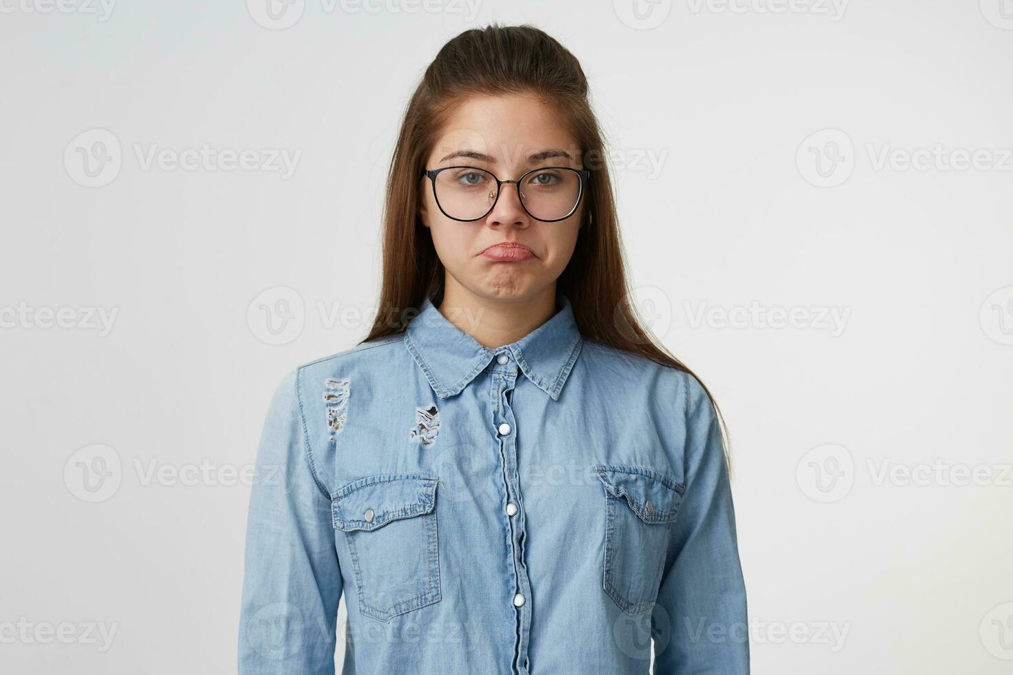 ritratto di un' giovane bella ragazza nel bicchieri con lungo capelli sembra triste irritato infelice imbronciato labbra, vestito nel un' denim camicia isolato su bianca sfondo foto