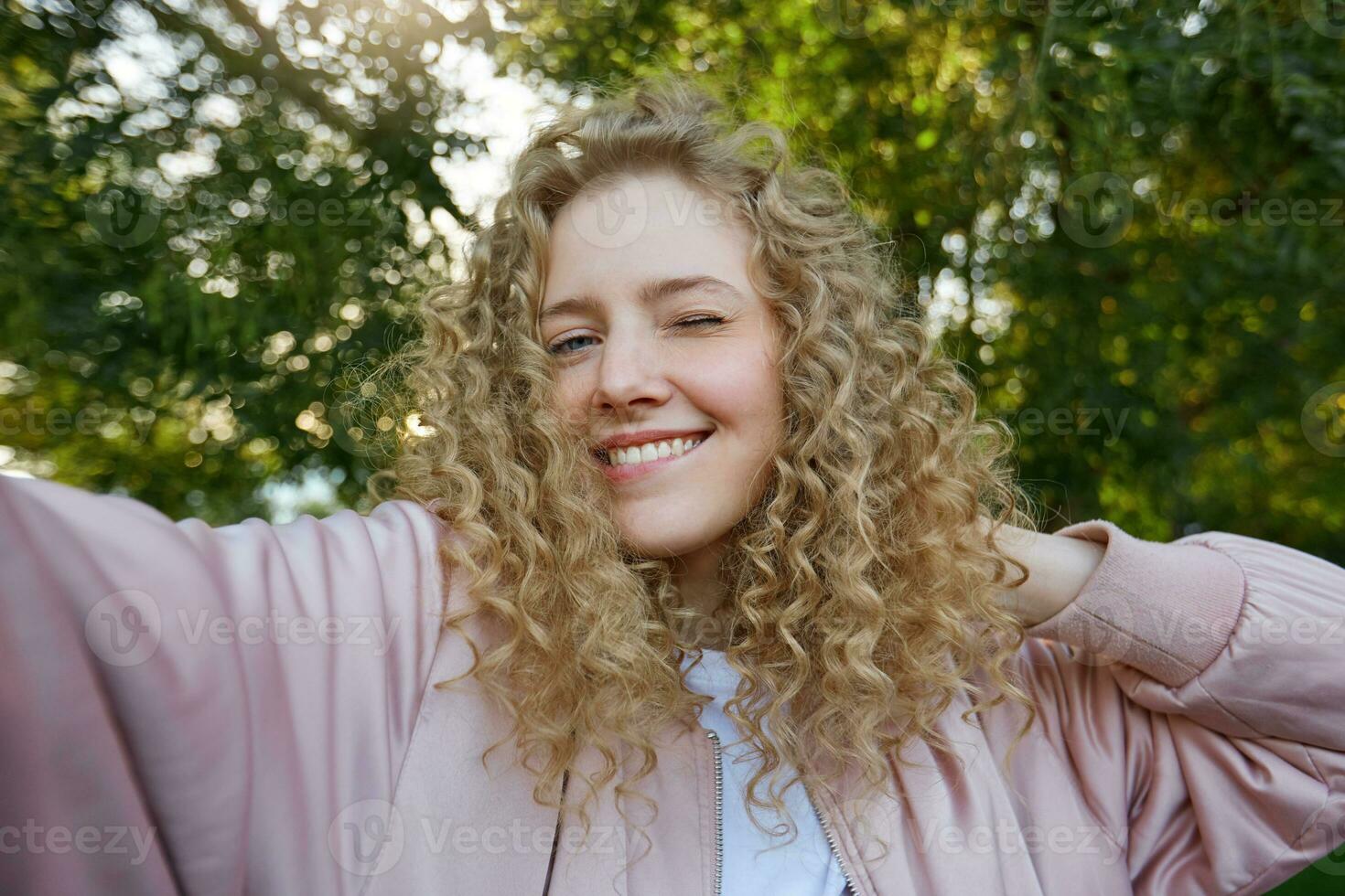 divertente attraente bella bionda ragazza con Riccio capelli, fabbricazione autoscatto su proprio Telefono per sociale reti, strizza l'occhio, po sua inferiore labbro, flirta, natura sfondo foto