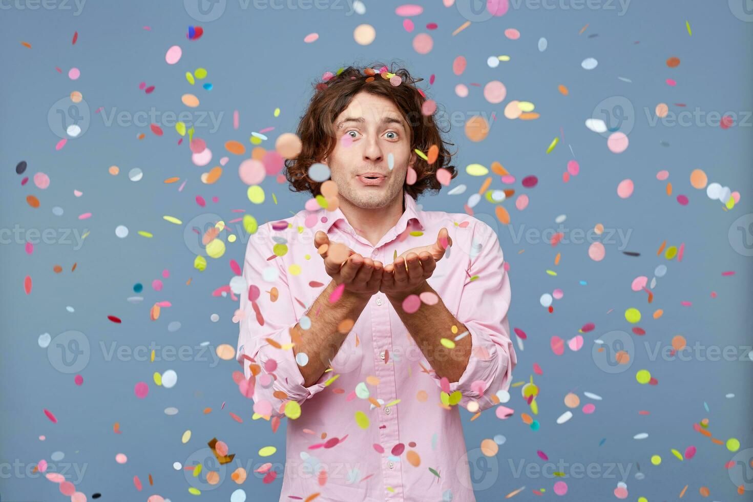 giovane uomo soffiaggio coriandoli via il suo mani. studio tiro di uomo contento in piedi in giro caduta giù coriandoli, al di sopra di blu sfondo foto