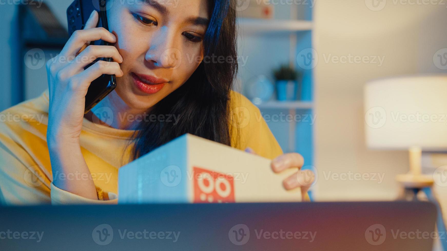 giovane donna asiatica chiama smartphone parla con il cliente per il controllo conferma l'ordine in magazzino sul computer portatile in ufficio a casa di notte. piccola impresa, consegna al mercato online, concetto di lifestyle freelance. foto