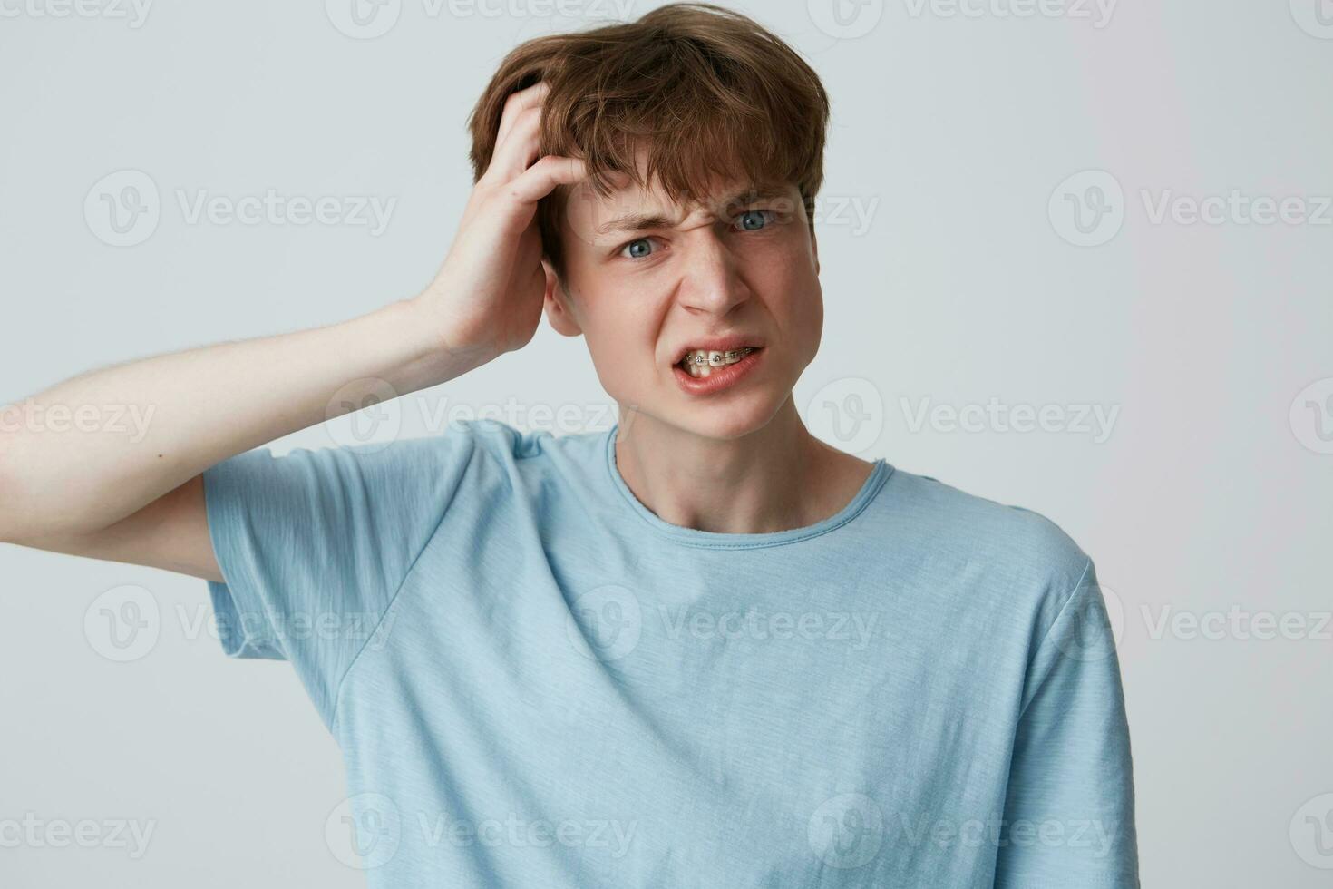 avvicinamento di arrabbiato pazzo giovane uomo con bretelle su denti indossa blu t camicia mantiene mano su testa e sembra irritata isolato al di sopra di bianca sfondo foto