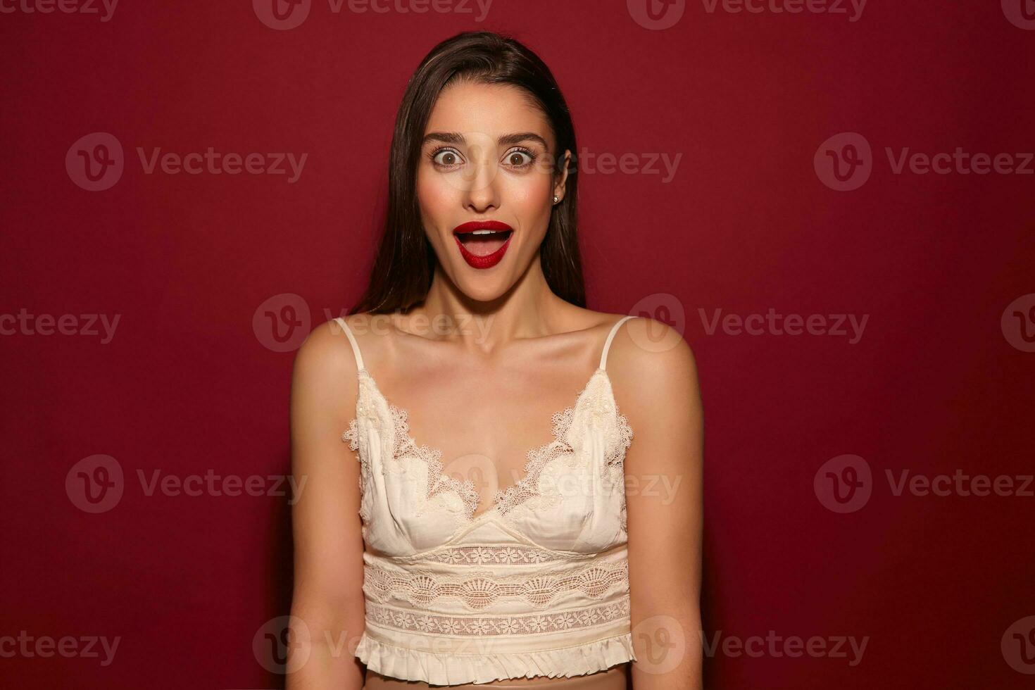 stupito giovane bello donna con buio lungo capelli guardare sorprendentemente a telecamera con largo occhi e bocca ha aperto, indossare festivo Abiti mentre in piedi al di sopra di Borgogna sfondo foto