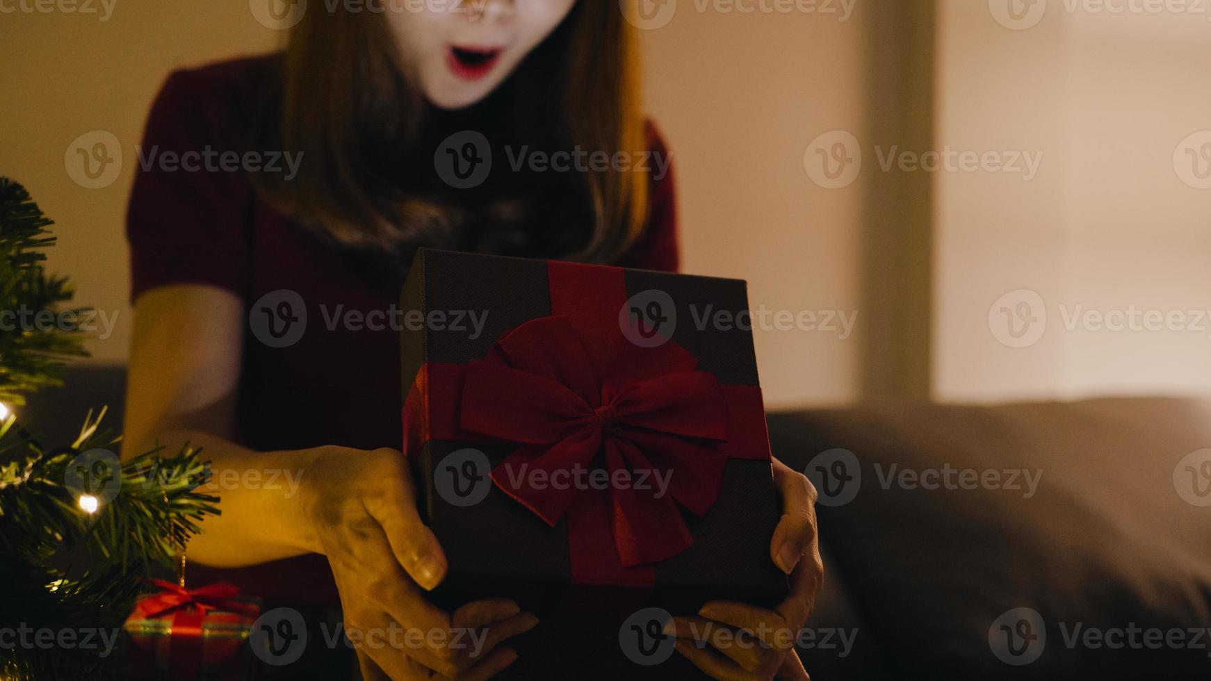 giovane donna asiatica che si diverte ad aprire la scatola regalo di Natale vicino all'albero di Natale decorato con ornamenti nel soggiorno di casa. buona notte di natale e felice anno nuovo festa delle vacanze. foto