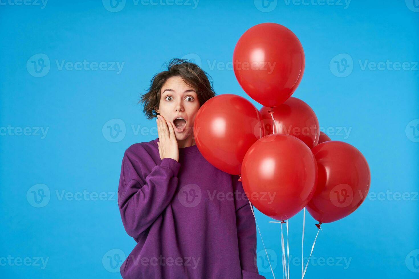 perplesso giovane bellissimo buio dai capelli donna conservazione palma su sua viso mentre guardare sorprendentemente a telecamera con ha aperto bocca, in posa al di sopra di blu sfondo con aria palloncini foto