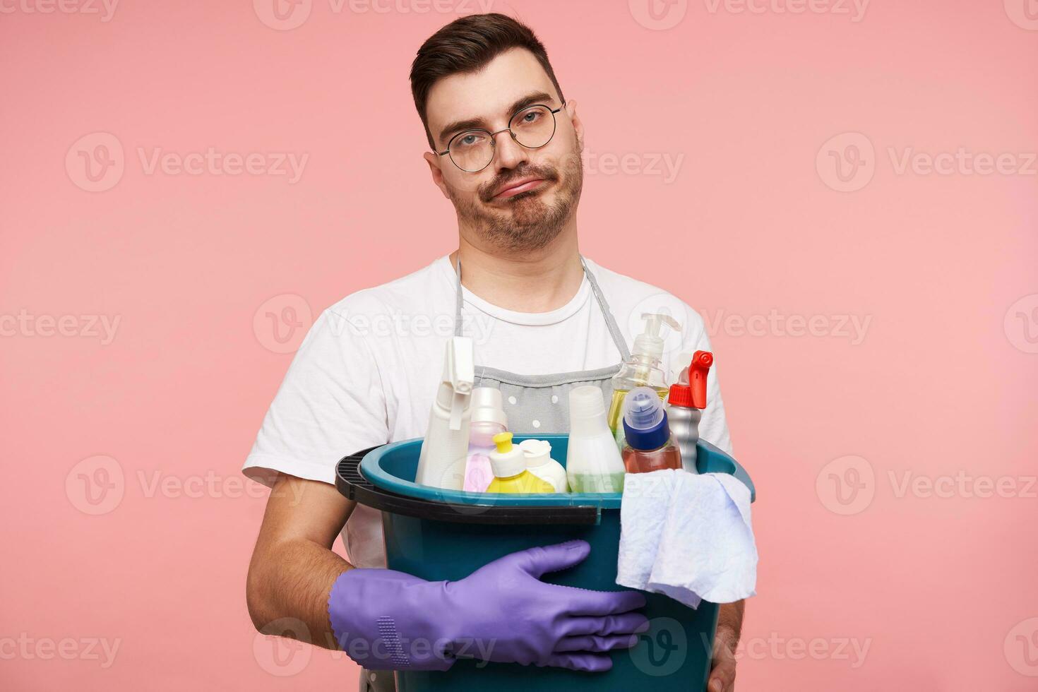 scontento giovane brunetta maschio con corto taglio di capelli smorfie il suo viso mentre conservazione busket con bottiglie di domestico sostanze chimiche, in piedi al di sopra di rosa sfondo nel uniforme foto