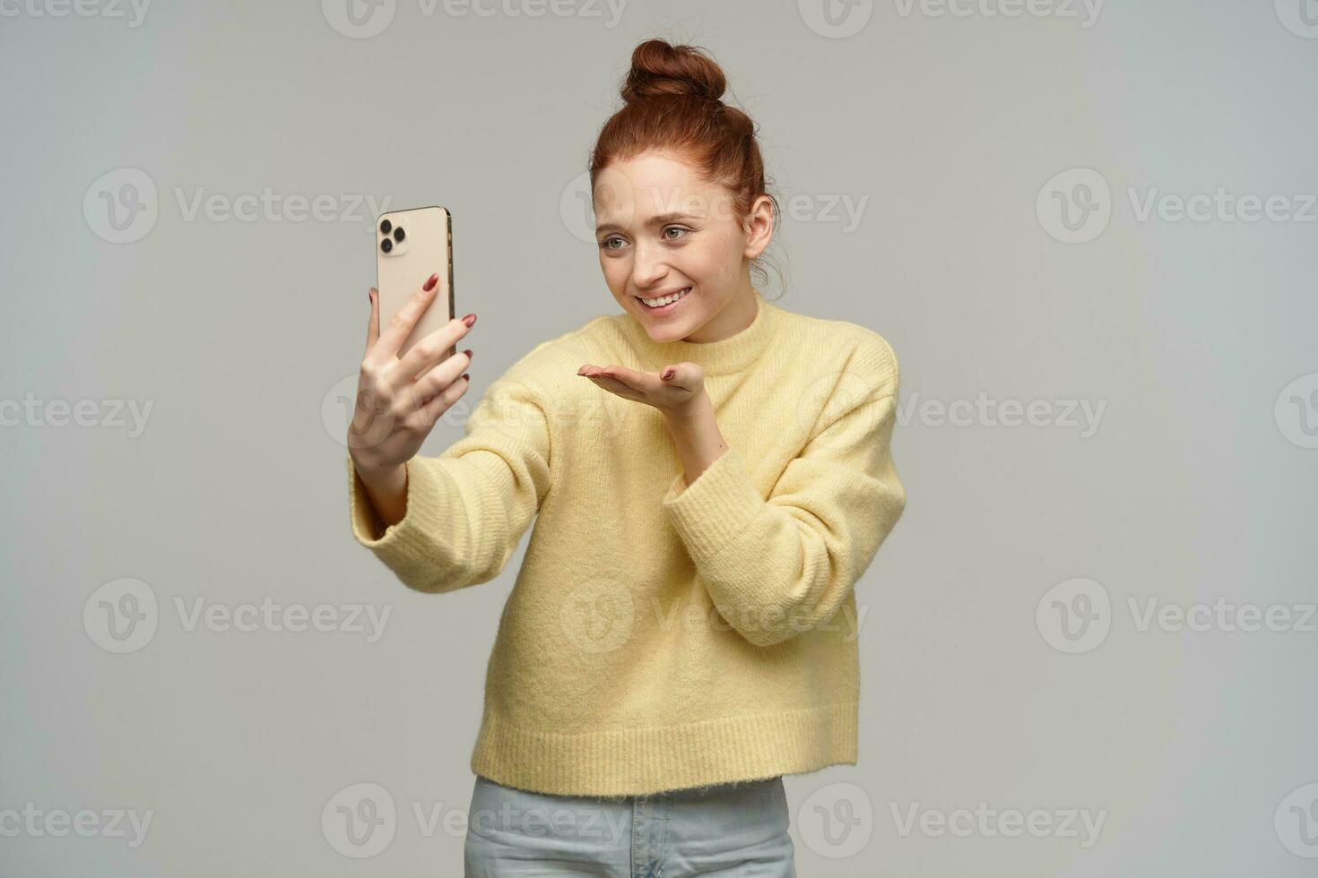 carino signora, bella donna con Zenzero capelli panino. indossare giallo maglione e Tenere un' smartphone. fabbricazione autoscatto, avere video chiamata. invio aria bacio, Sorridi. In piedi isolato al di sopra di grigio sfondo foto