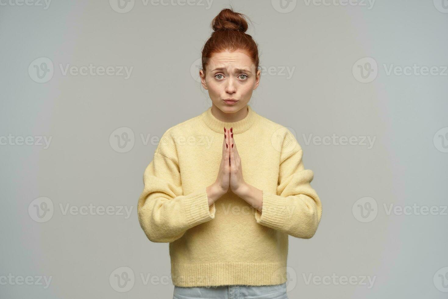 triste donna con Zenzero capelli si riunirono nel un' panino. indossare pastello giallo maglione e jeans. mantenere sua palme insieme nel preghiera. Chiedi per qualche cosa. Guardando a il telecamera isolato al di sopra di grigio sfondo foto