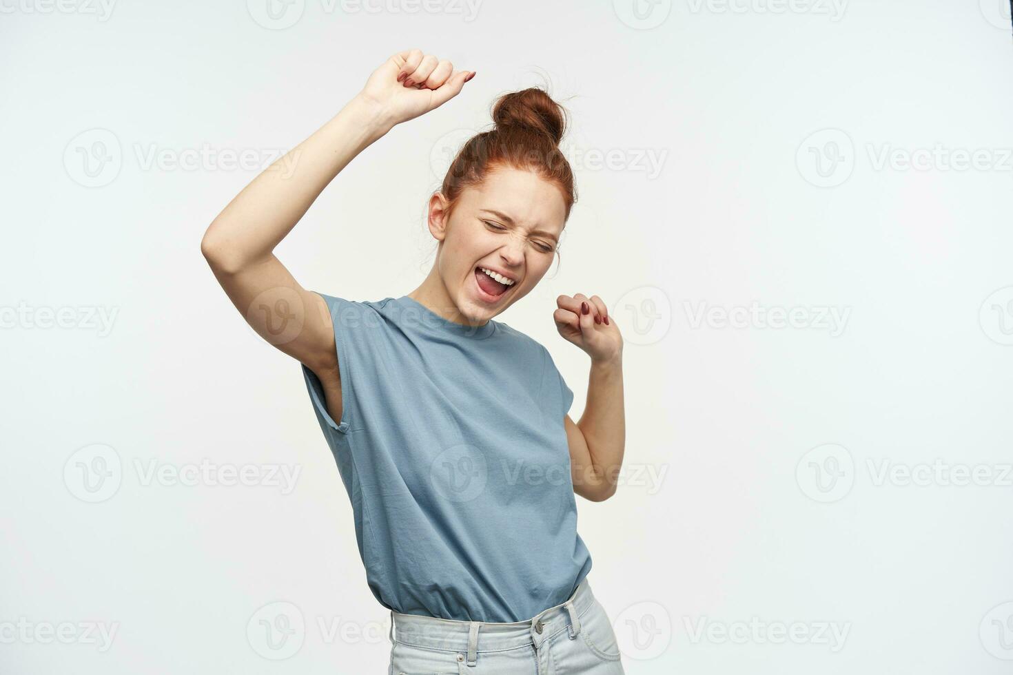 ritratto di attraente, contento testa Rossa ragazza con capelli si riunirono nel un' panino. indossare blu maglietta e jeans. emozionato. ascensori sua braccia e danza con chiuso occhi. In piedi isolato al di sopra di bianca sfondo foto