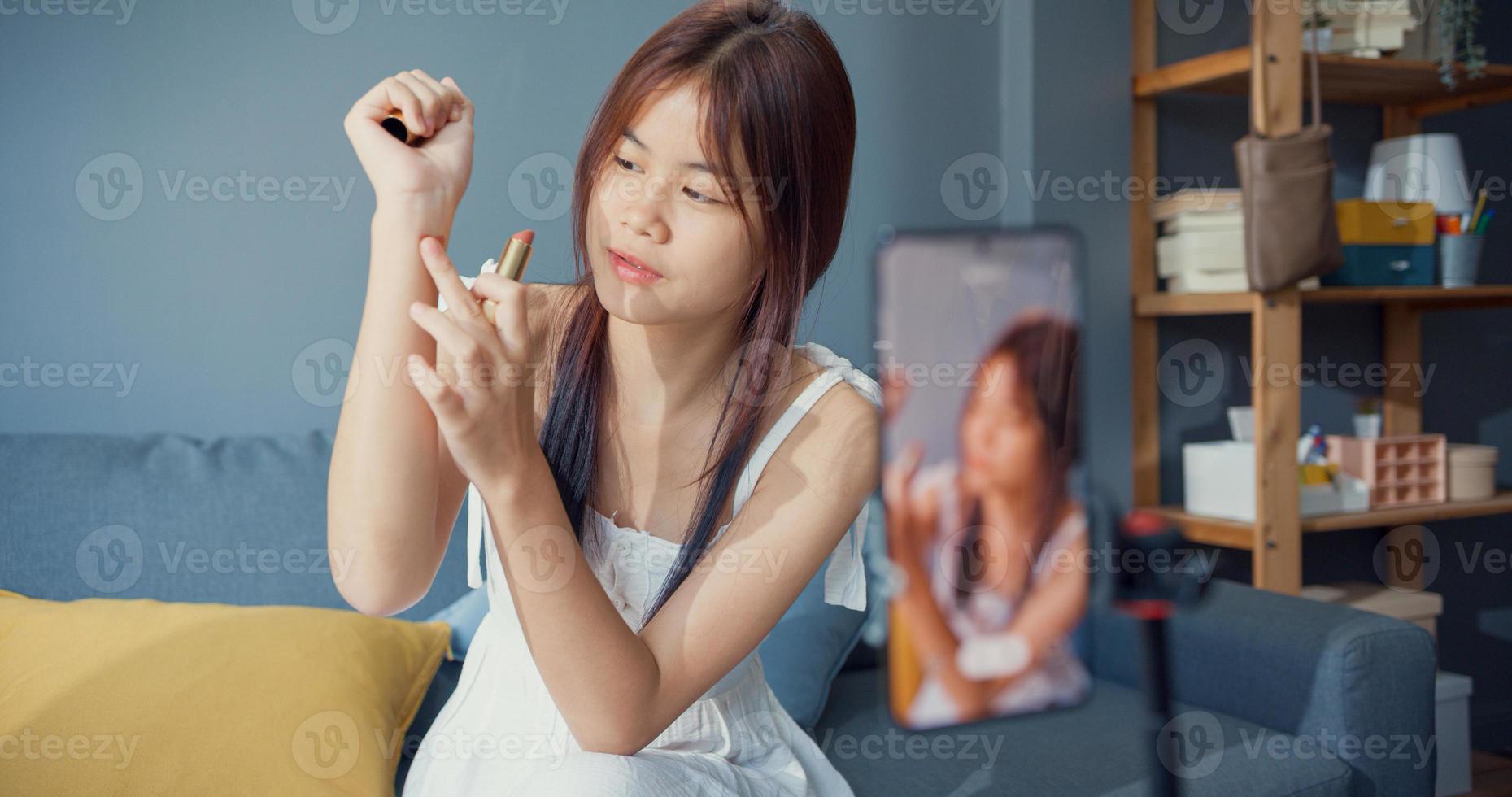 felice giovane ragazza asiatica trucco vlog anteriore della fotocamera del telefono godere di revisione rossetto parlare con seguace nel soggiorno a casa. stile di vita dell'attività di blogger, concetto di pandemia di coronavirus a distanza sociale. foto