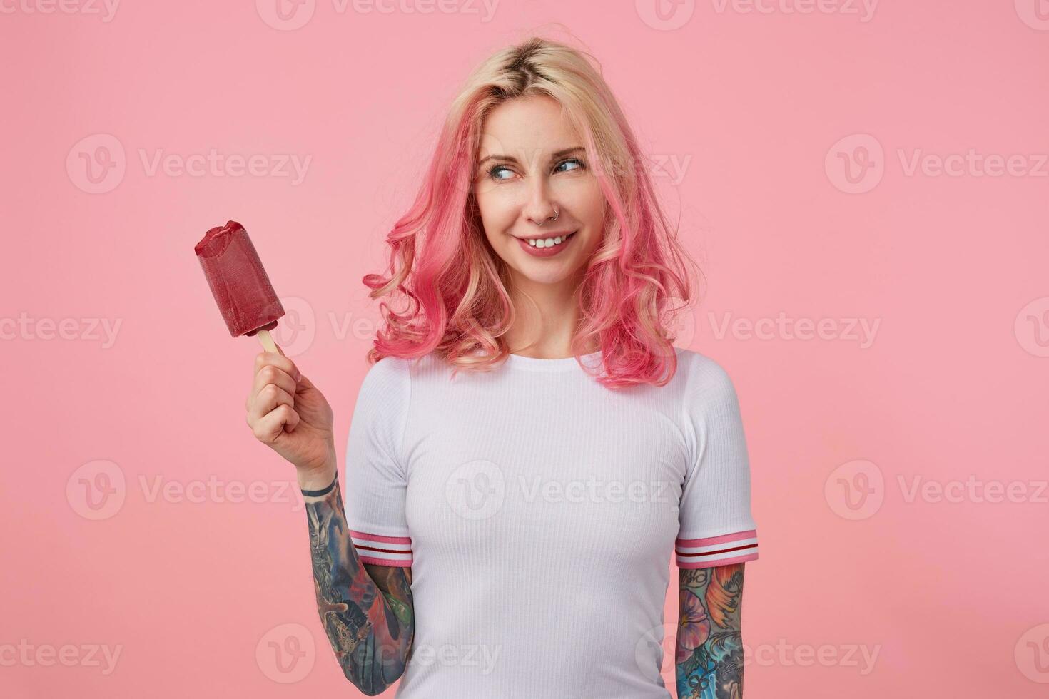 ritratto di contento carino ragazza con rosa capelli e tatuato mani, guardare a ghiaccio crema nel il suo sinistra mano e sorridente, in piedi al di sopra di rosa sfondo, indossare un' bianca maglietta. foto