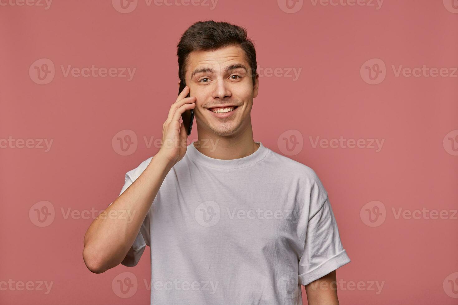 giovane bello allegro uomo indossa nel vuoto maglietta, sembra a il telecamera con contento espressione, sta al di sopra di rosa sfondo e parla con amico su il telefono. foto