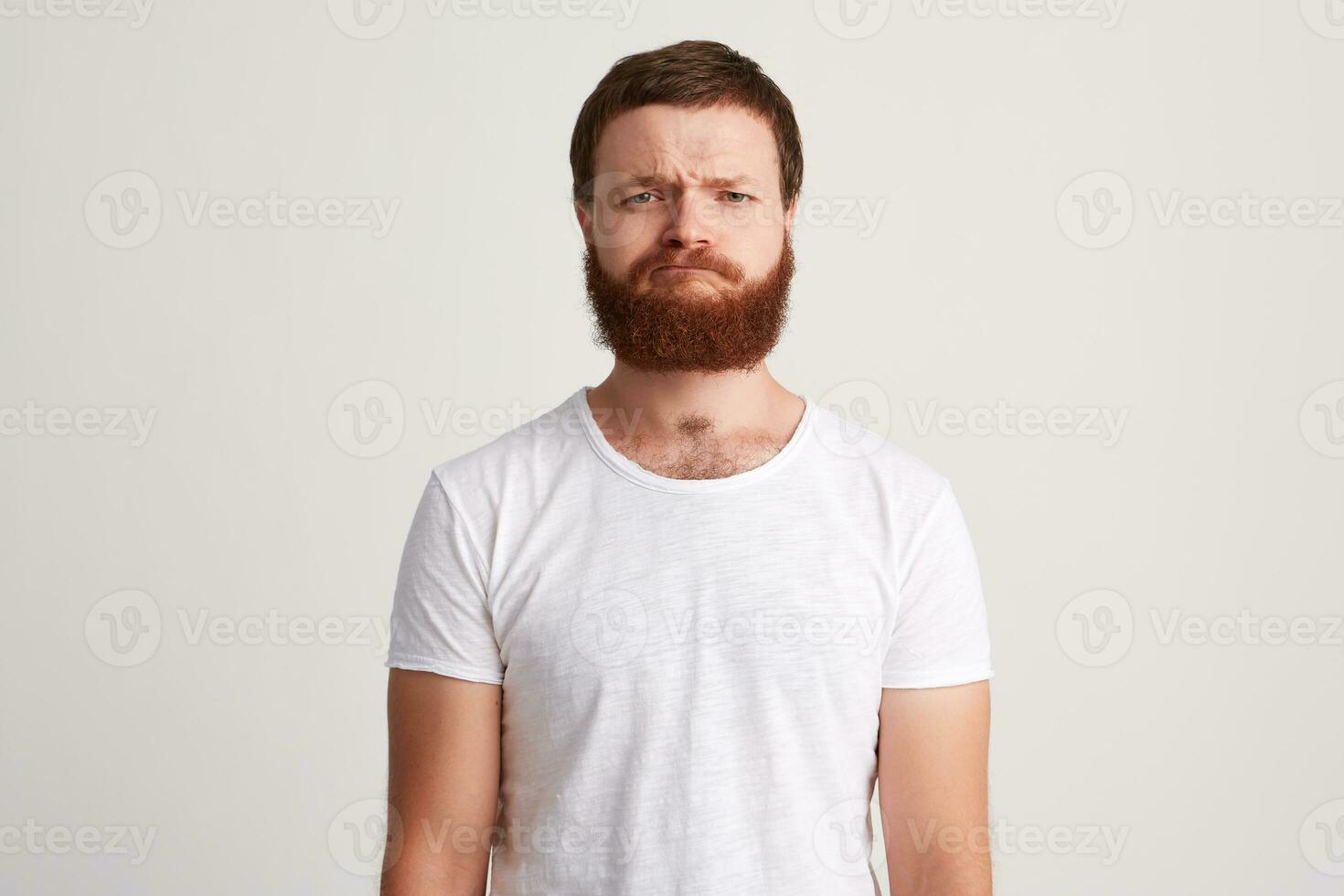 ritratto di grave rigoroso barbuto giovane uomo indossa t camicia in piedi e si sente preoccupato isolato al di sopra di bianca sfondo sembra direttamente nel telecamera foto