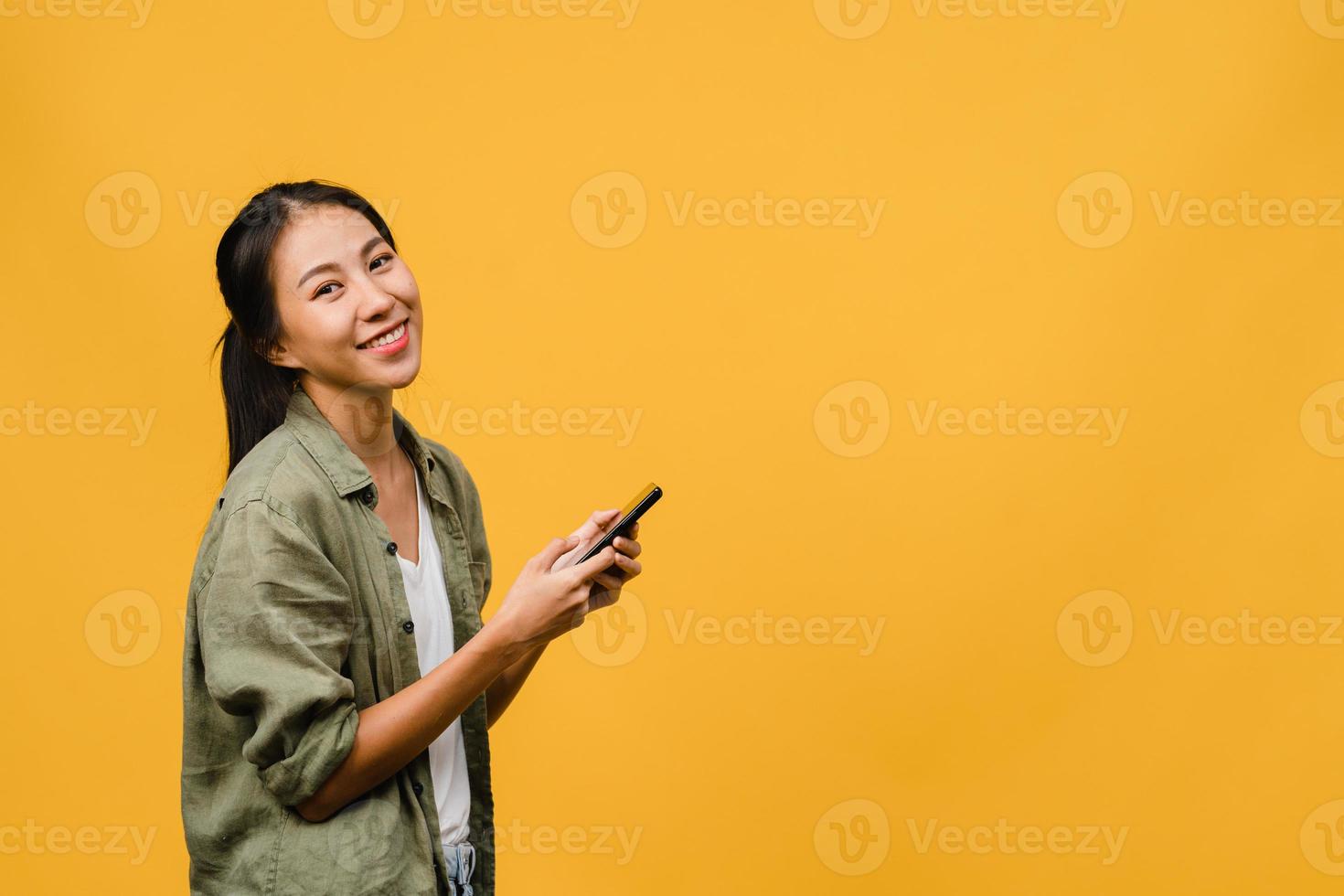 sorpresa giovane signora asiatica che utilizza il telefono cellulare con espressione positiva, sorride ampiamente, vestita con abiti casual e guardando la telecamera su sfondo giallo. felice adorabile donna felice esulta successo. foto