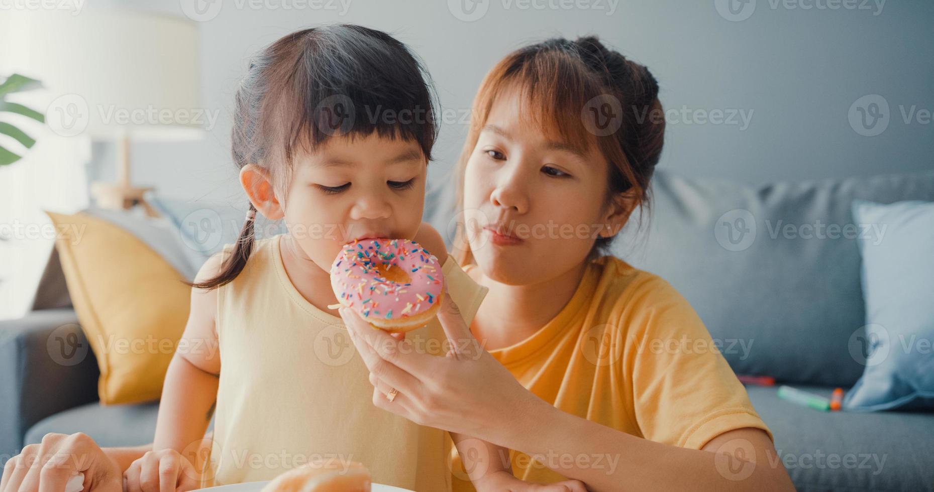 felice allegra famiglia asia mamma e bambina che mangiano ciambelle e si divertono rilassati goditi sul divano nel soggiorno di casa. passare del tempo insieme, distanza sociale, quarantena per il coronavirus. foto