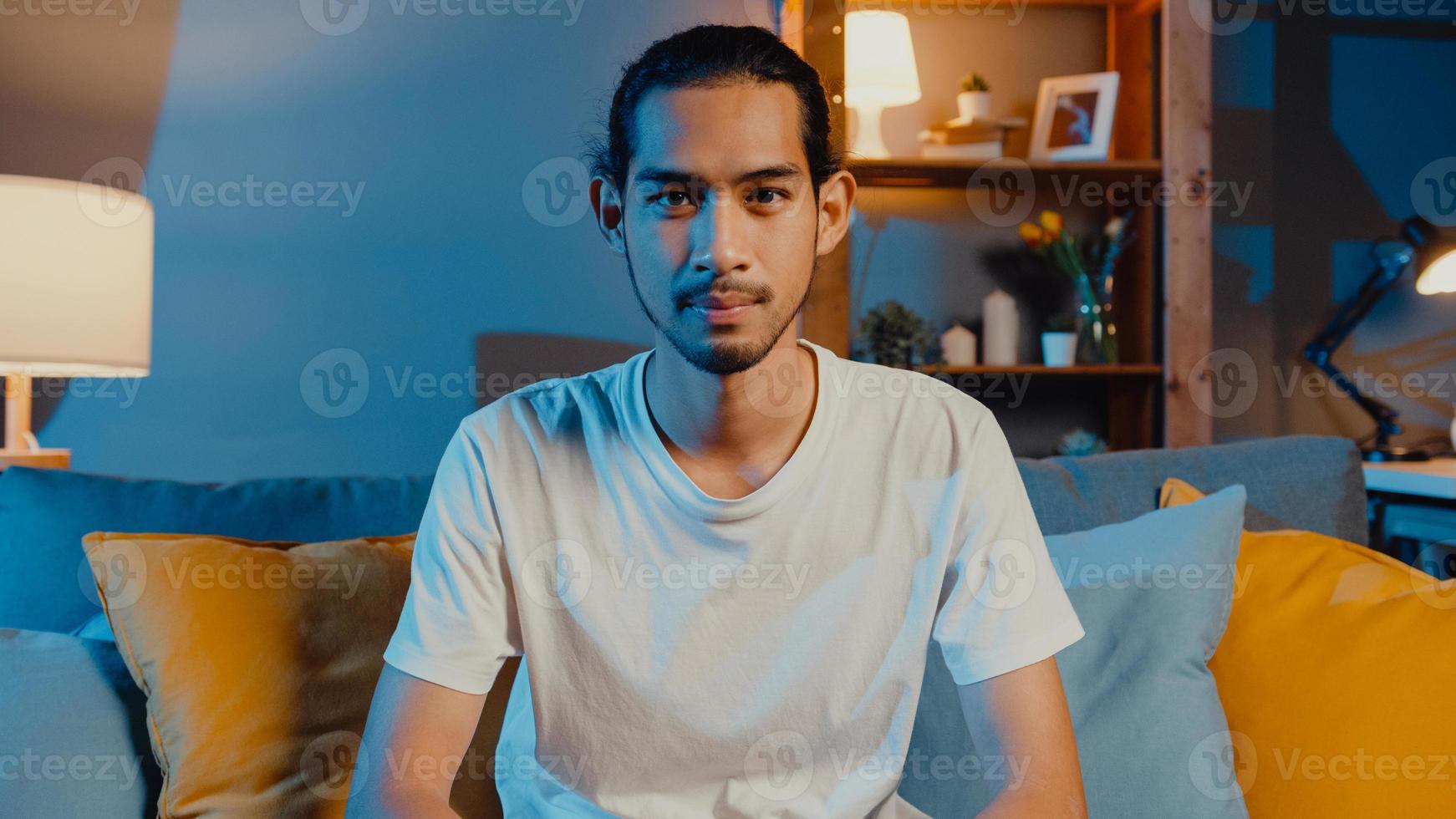 felice giovane uomo asiatico freelance che guarda l'obbiettivo sorridente e allegro rilassarsi durante la videochiamata online di notte nel soggiorno di casa, stare a casa in quarantena, lavorare da casa, concetto di distanza sociale. foto