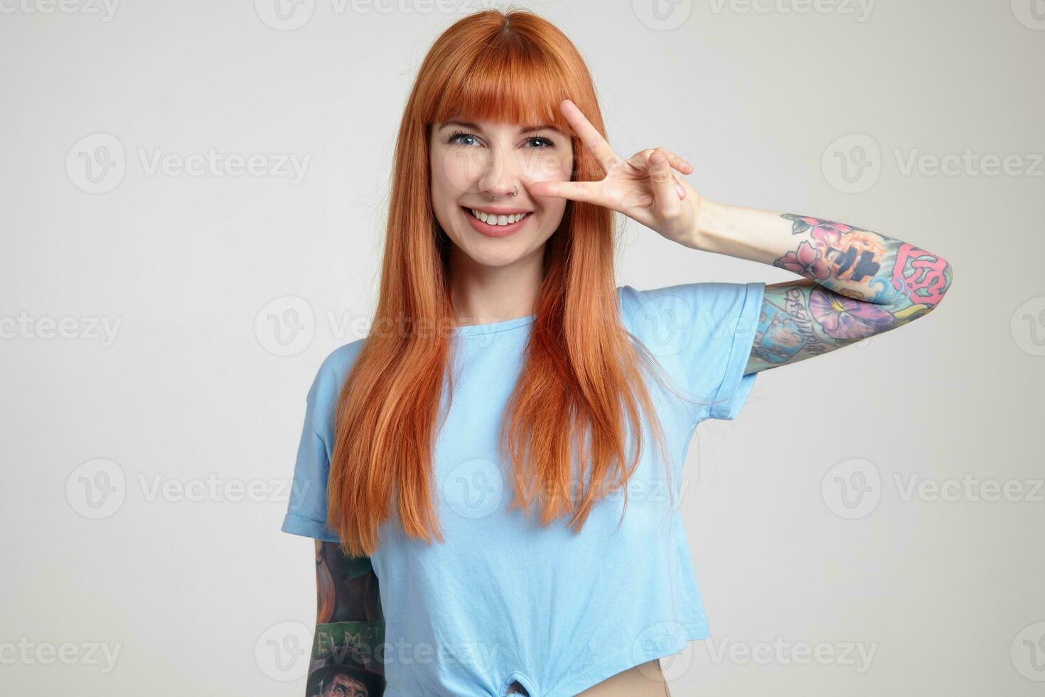 interno ritratto di giovane Zenzero femmina in posa al di sopra di bianca parete mostrando vittoria cartello in telecamera e Sorridi a grandi linee foto