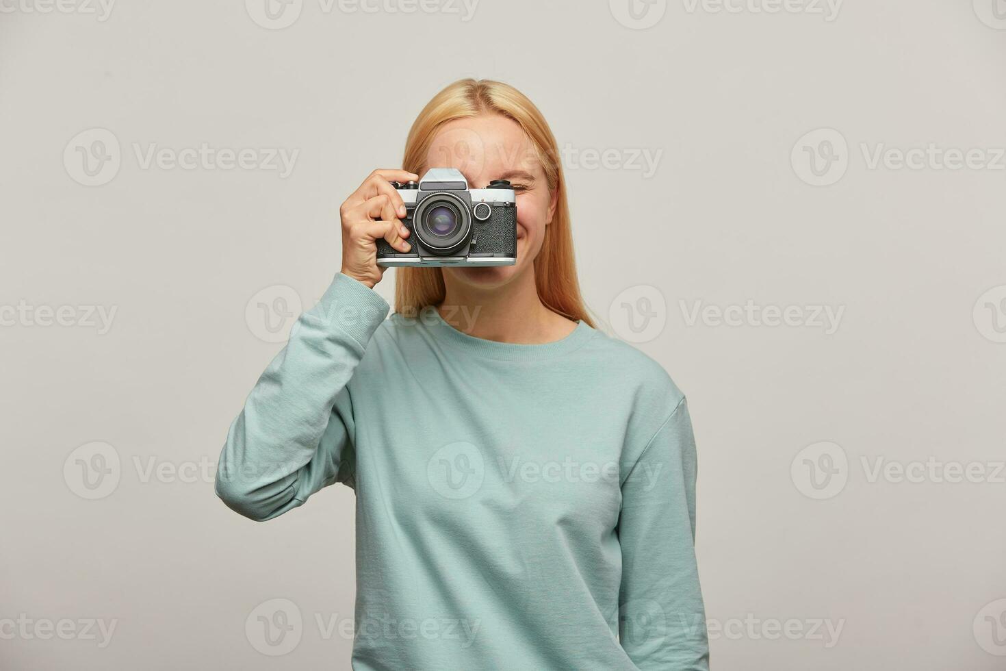 vicino su di un' fotografo copertura sua viso con il retrò Vintage ▾ telecamera. contento con il telecamera, piace per prendere immagini, -Tenuto in mano fotografia, vestito nel blu felpa, al di sopra di grigio sfondo foto