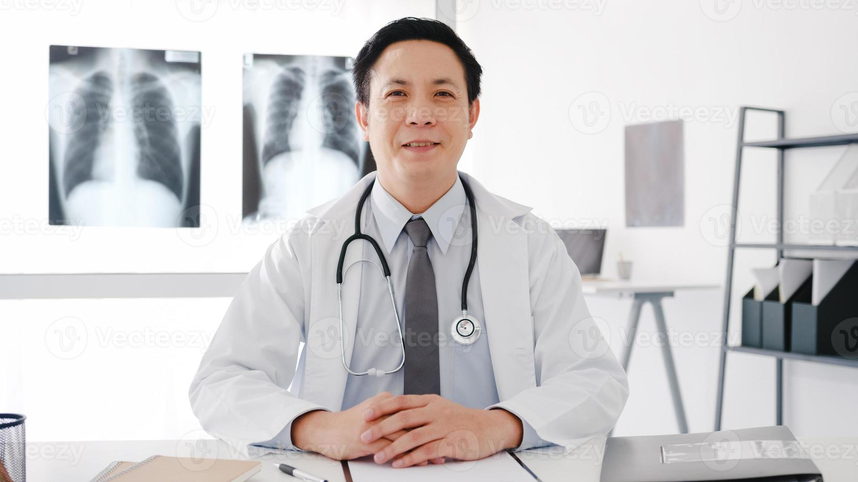 fiducioso giovane medico maschio asiatico in uniforme medica bianca con stetoscopio che guarda l'obbiettivo e sorride durante la videoconferenza con il paziente in ospedale. concetto di consulenza e terapia. foto