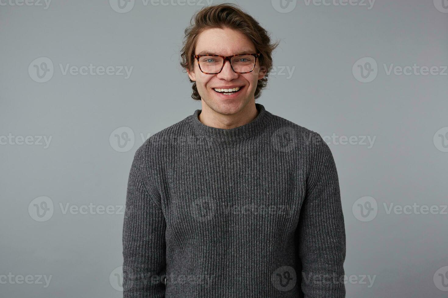 foto di giovane positivo bello uomo con bicchieri indossa nel grigio maglione, sta al di sopra di grigio sfondo e a grandi linee sorrisi, sembra contento e allegro.