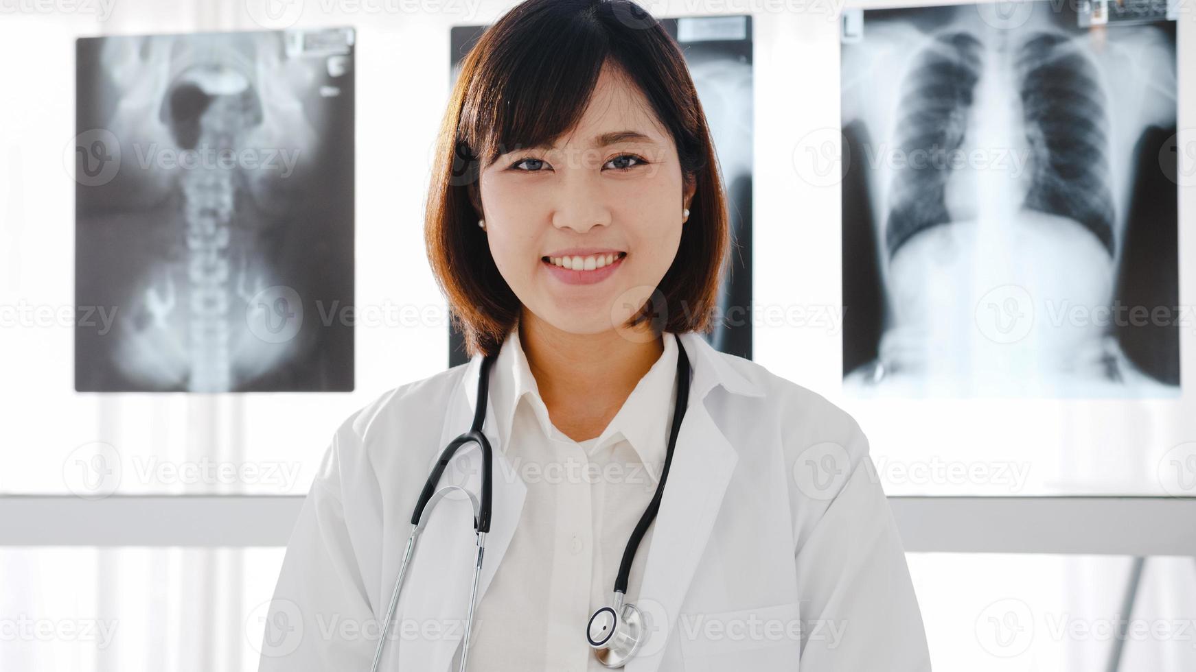 fiducioso giovane dottoressa asiatica in uniforme medica bianca con stetoscopio che guarda l'obbiettivo e sorride durante la videoconferenza con il paziente in ospedale. concetto di consulenza e terapia. foto