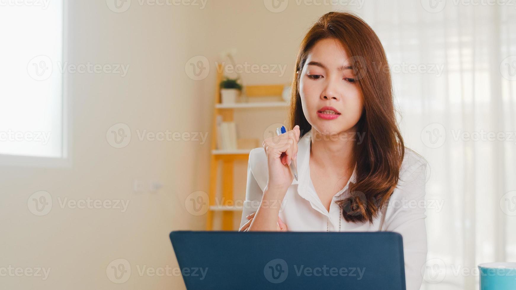 donna d'affari asiatica che utilizza laptop parla con i colleghi del piano in videochiamata mentre lavora in modo intelligente da casa in soggiorno. autoisolamento, distanziamento sociale, quarantena per la prevenzione del coronavirus. foto