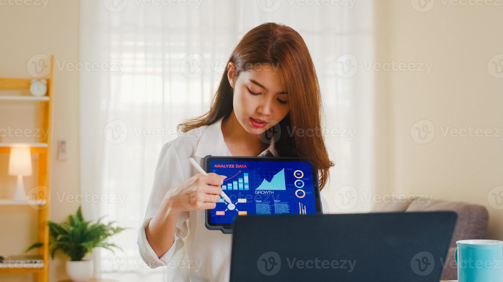 donna d'affari asiatica che utilizza laptop e tablet presentazione ai colleghi sul piano in videochiamata mentre si lavora da casa in soggiorno. autoisolamento, distanziamento sociale, quarantena per coronavirus. foto