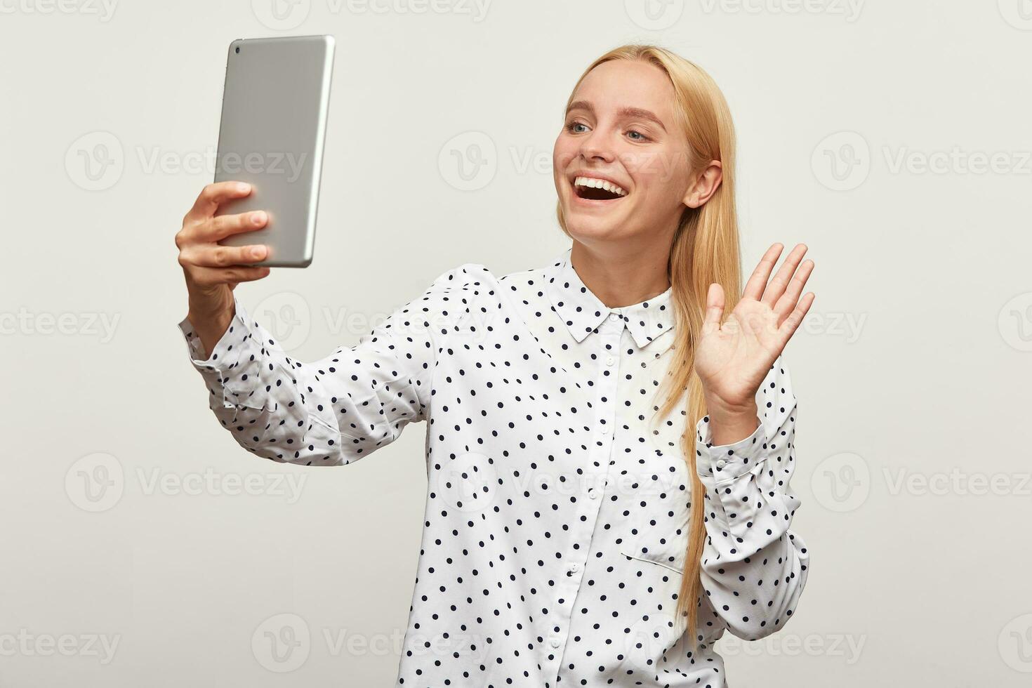 sparare di un' bionda giovane ragazza con tavoletta nel mano, fabbricazione un' video chiamata o registrazione video, sorridente agitando guardare tavoletta schermo, indossa bianca camicia con nero punti, isolato al di sopra di bianca sfondo foto