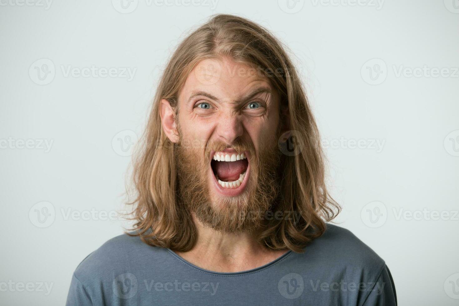 ritratto di isterico irritata barbuto giovane uomo con bionda lungo capelli indossa grigio t camicia sembra pazzo e urlando isolato al di sopra di bianca sfondo foto
