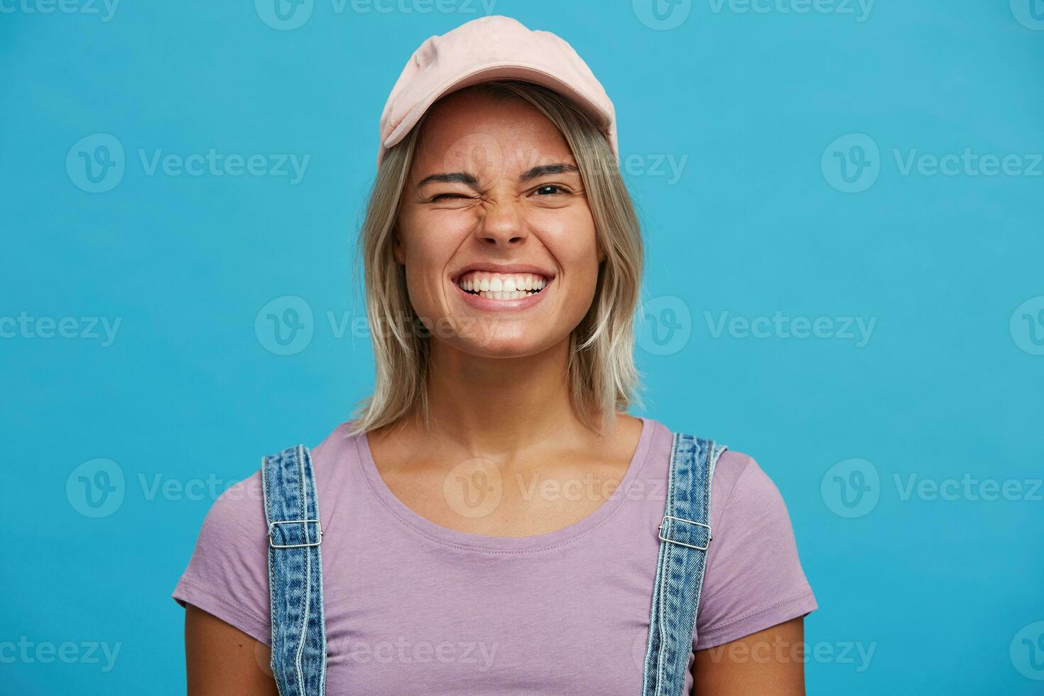 avvicinamento di allegro bellissimo bionda giovane donna indossa rosa berretto e viola t camicia sembra felice, sorridente e strizzando l'occhio isolato al di sopra di blu sfondo foto