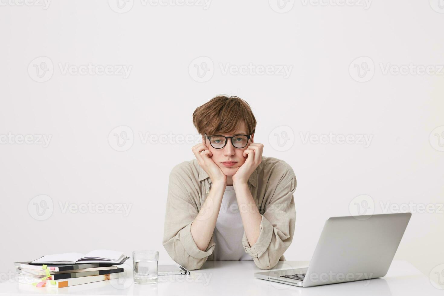 avvicinamento di infelice stanco giovane uomo alunno indossa bicchieri e beige camicia sembra triste e Lavorando con il computer portatile computer e i Quaderni a il tavolo isolato al di sopra di bianca sfondo foto