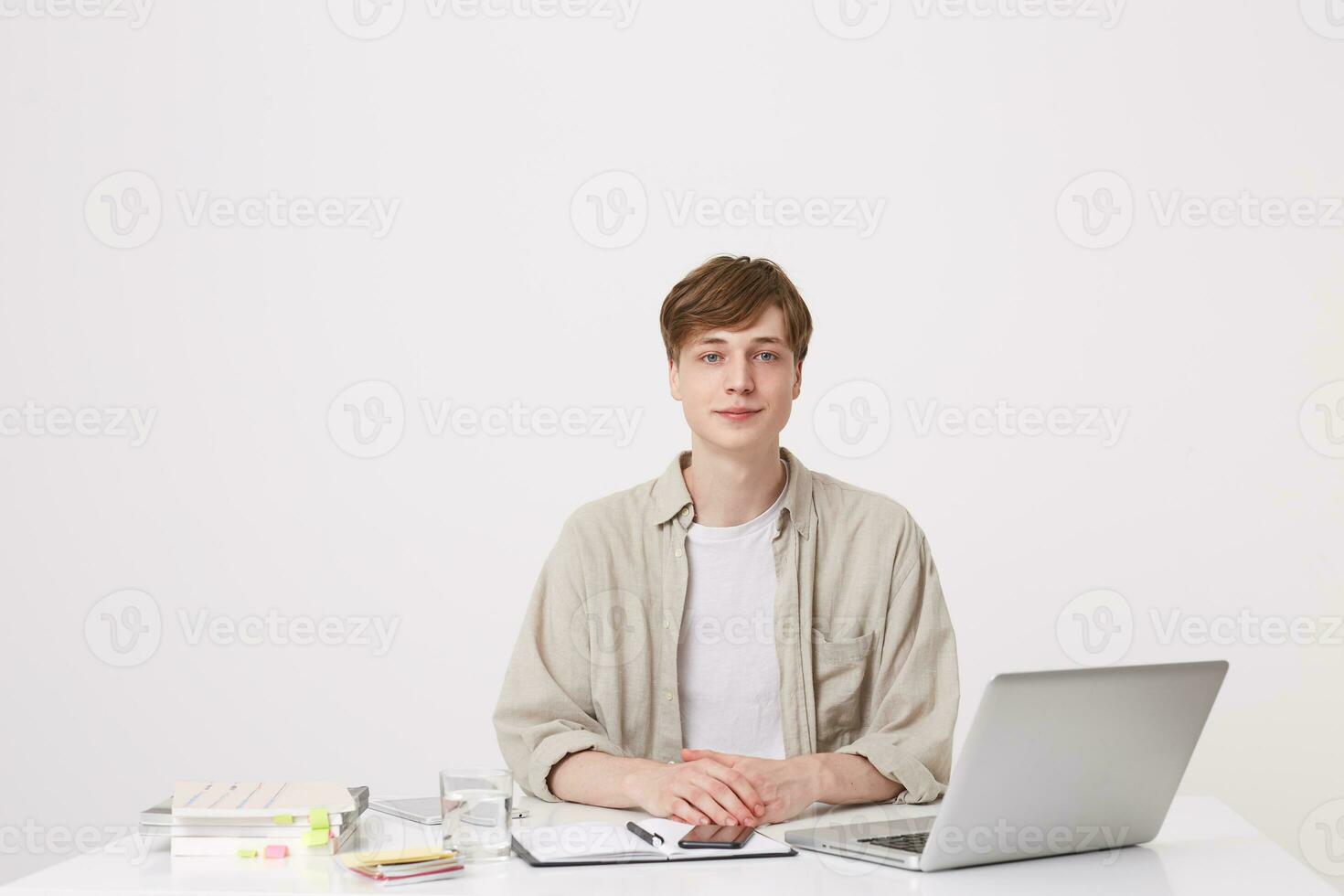 avvicinamento di fiducioso giovane uomo alunno indossa beige camicia seduta e Lavorando con il computer portatile computer e i Quaderni a il tavolo isolato al di sopra di bianca sfondo foto