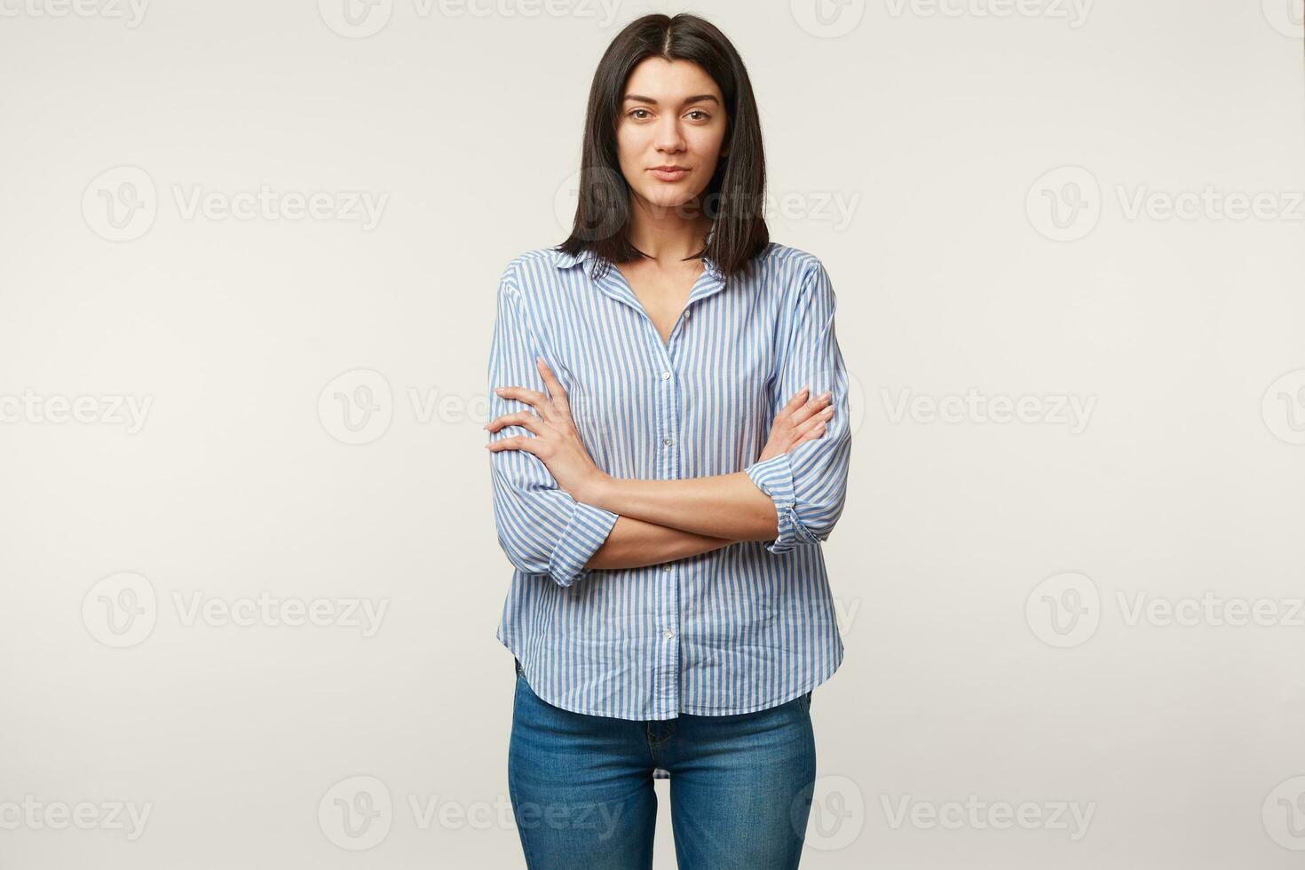 giovane brunetta donna, calma, attento, ascolta con rispetto, paga Attenzione, in piedi con braccia attraversato vestito nel blu jeans e a strisce camicia isolato al di sopra di bianca sfondo foto