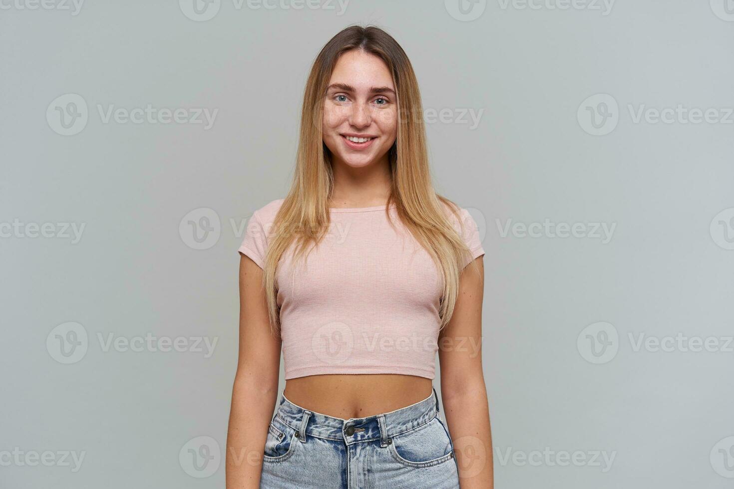 adolescenziale ragazza, contento guardare donna con biondo lungo capelli. indossare rosa maglietta e jeans. persone e emozione concetto. Guardando con Sorridi a il telecamera isolato al di sopra di grigio sfondo foto