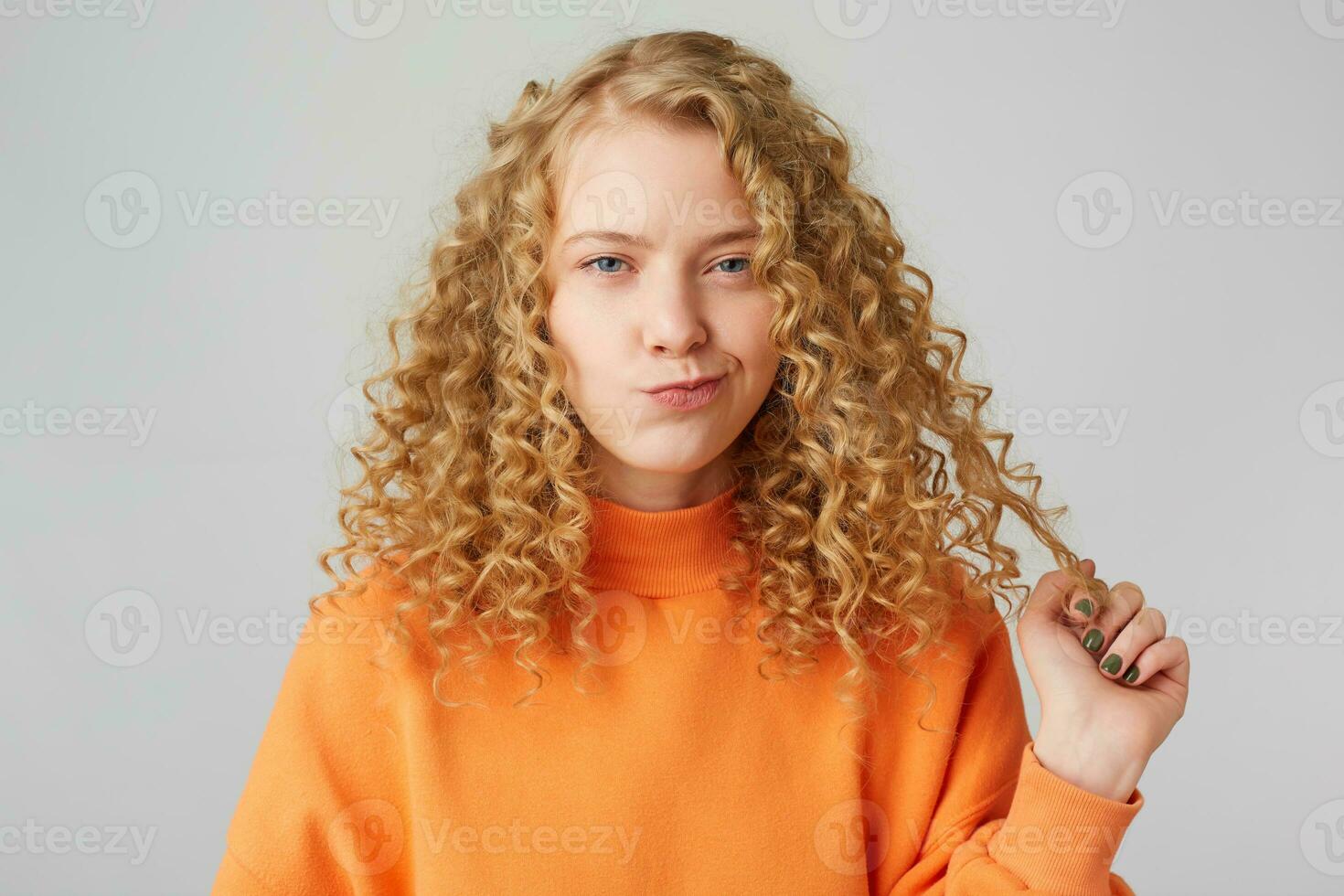 carino dai capelli ricci bionda con blu occhi sospettosamente meticolosamente attentamente incredulo guardare a il fotocamera, tiene un' filo di capelli pensiero di qualcosa, dentro arancia maglione, passo bianca sfondo. foto