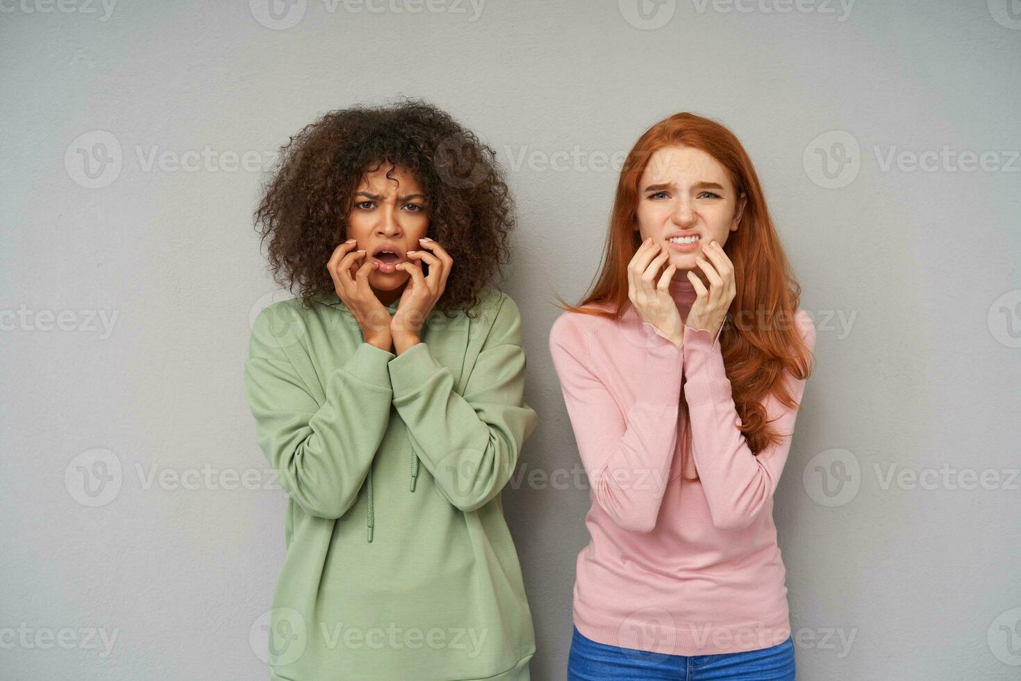 ritratto di confuso giovane bella amiche accigliato loro sopracciglia e Tenere sollevato mani su guance mentre guardare a telecamera con broncio, in piedi contro grigio sfondo foto