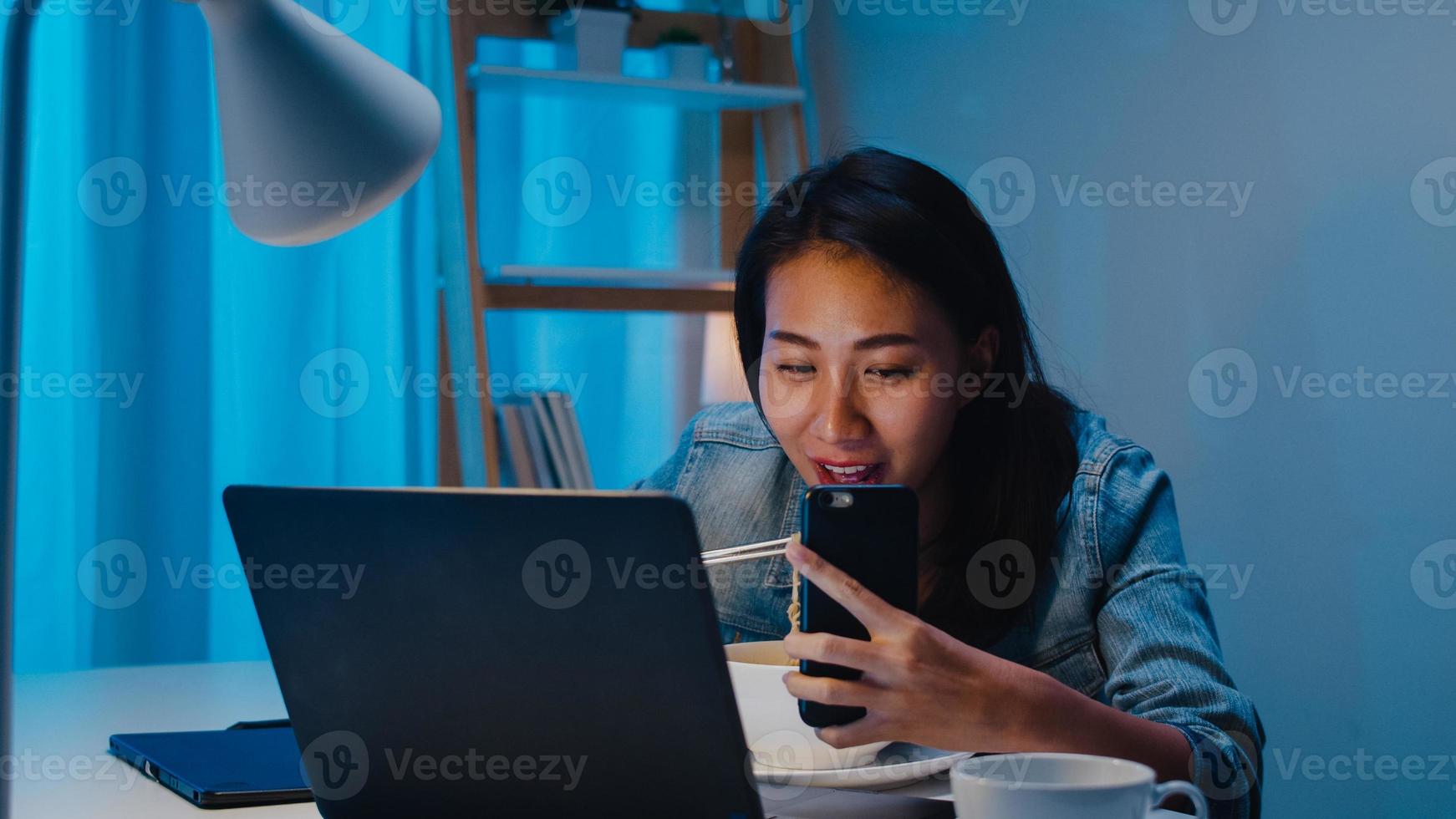 donne d'affari intelligenti freelance che mangiano noodles istantanei in diretta sui social media tramite telefono cellulare nel soggiorno di casa di notte. la giovane ragazza asiatica felice che si siede sulla scrivania fa gli straordinari, godetevi il tempo di relax. foto