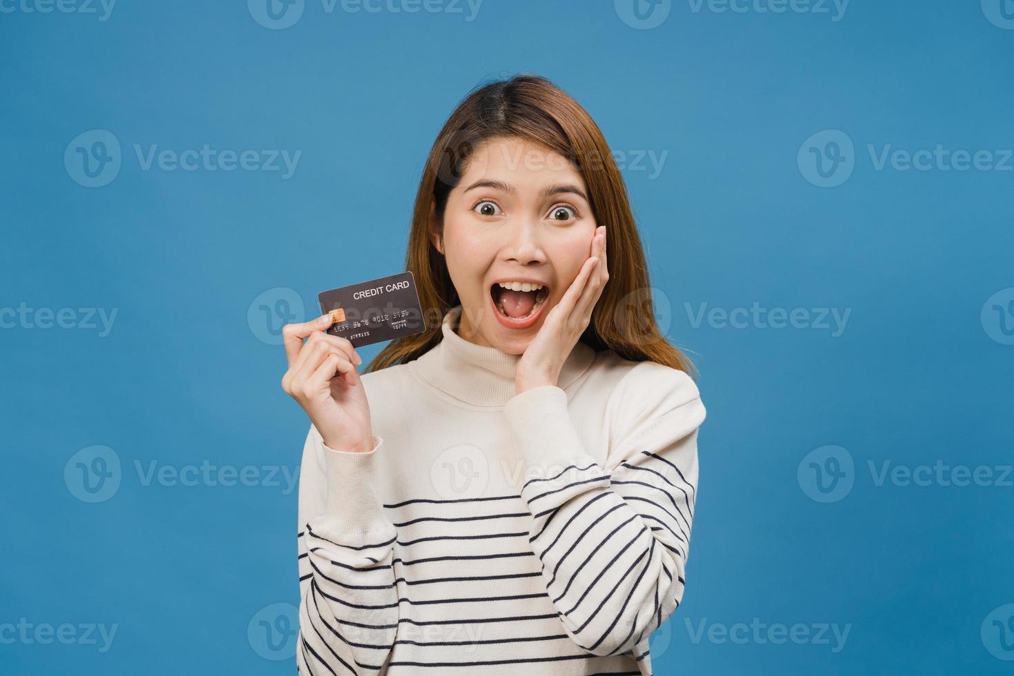 la giovane donna asiatica mostra la carta di credito con l'espressione positiva, sorride ampiamente, vestita con abiti casual sentendo felicità e stando isolata su sfondo blu. concetto di espressione facciale. foto