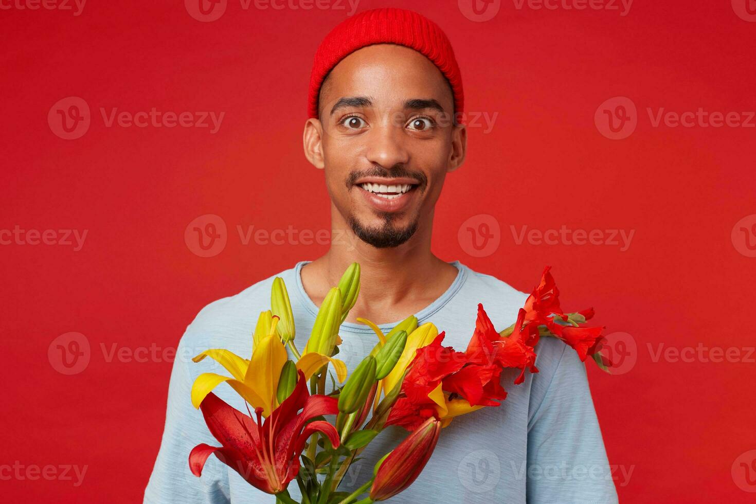 ritratto di giovane allegro attraente tipo nel rosso cappello e blu maglietta, detiene un' mazzo nel il suo mani, sembra a il telecamera con contento espressione e sorridente, sta al di sopra di rosso sfondo. foto