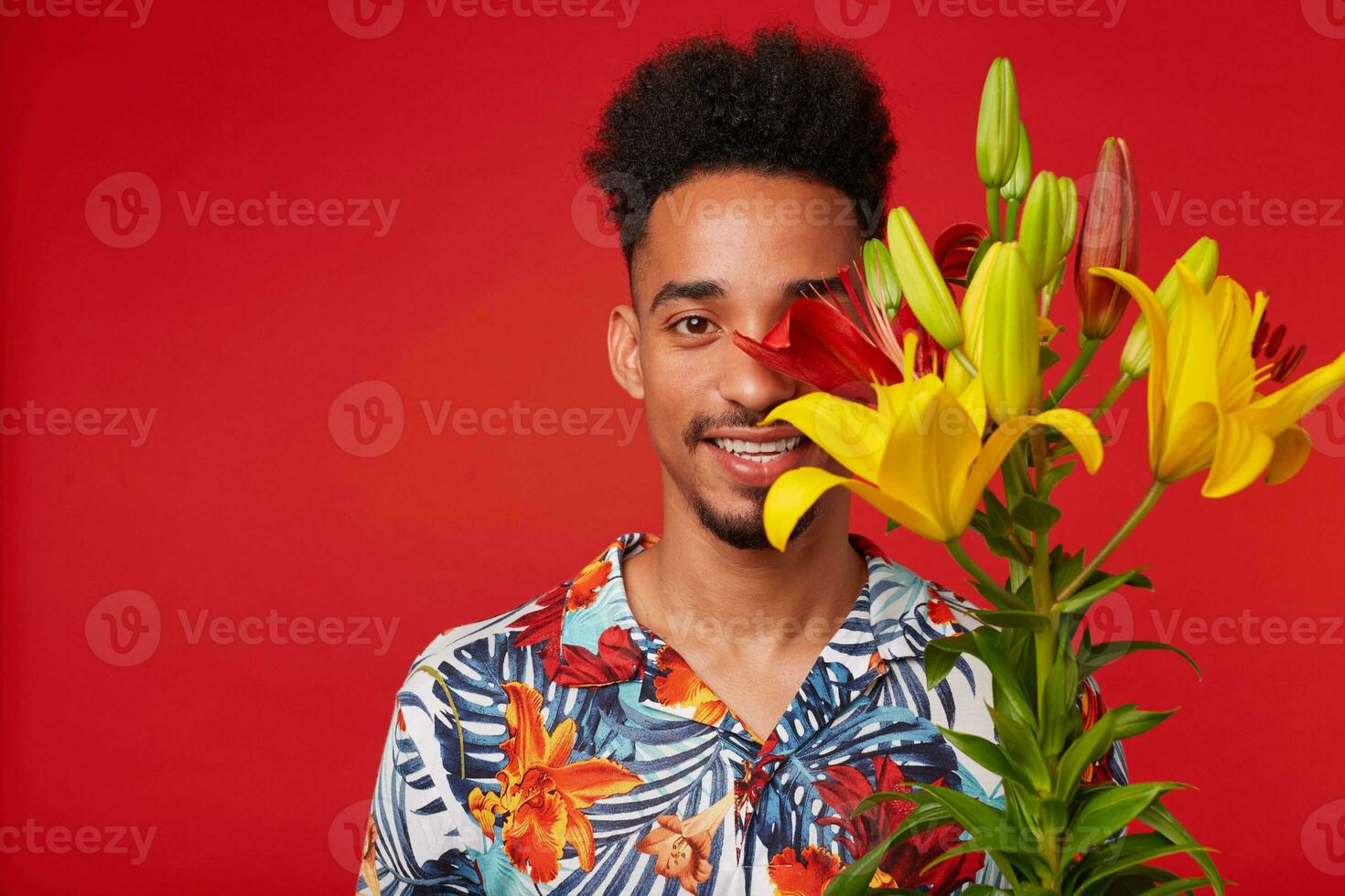 vicino su di giovane contento africano americano uomo, indossa nel hawaiano camicia, sembra a il telecamera con contento espressione, detiene giallo e rosso fiori e coperto viso, sta al di sopra di rosso sfondo. foto