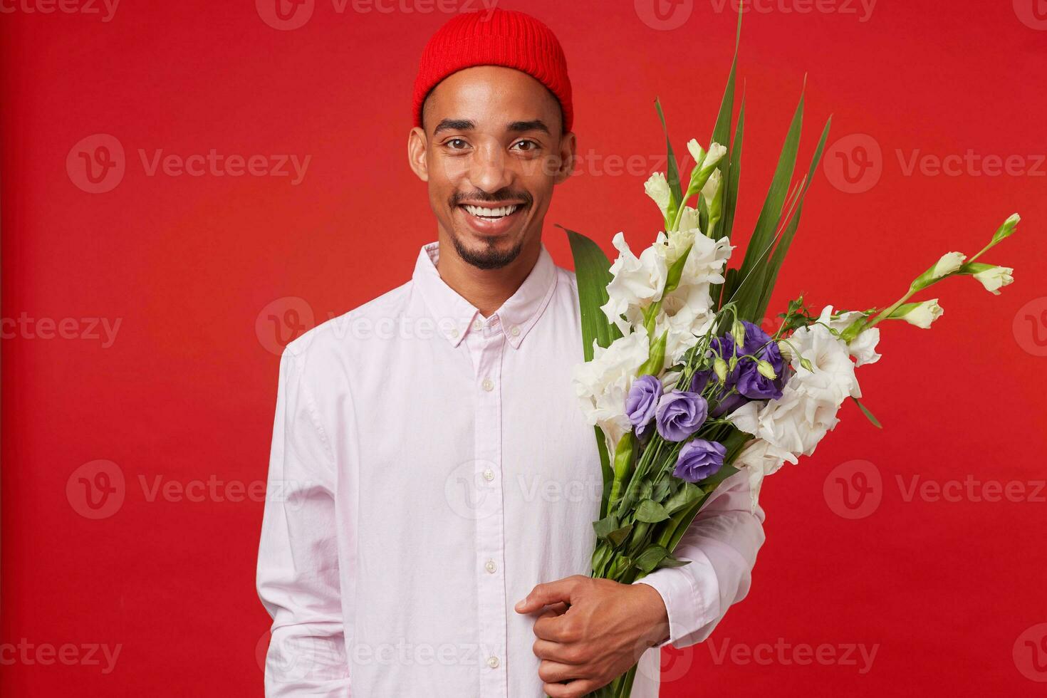 giovane positivo africano americano attraente tipo, indossa nel bianca camicia e rosso cappello, sembra a il telecamera e detiene mazzo , sta al di sopra di rosso sfondo e sorridente. foto