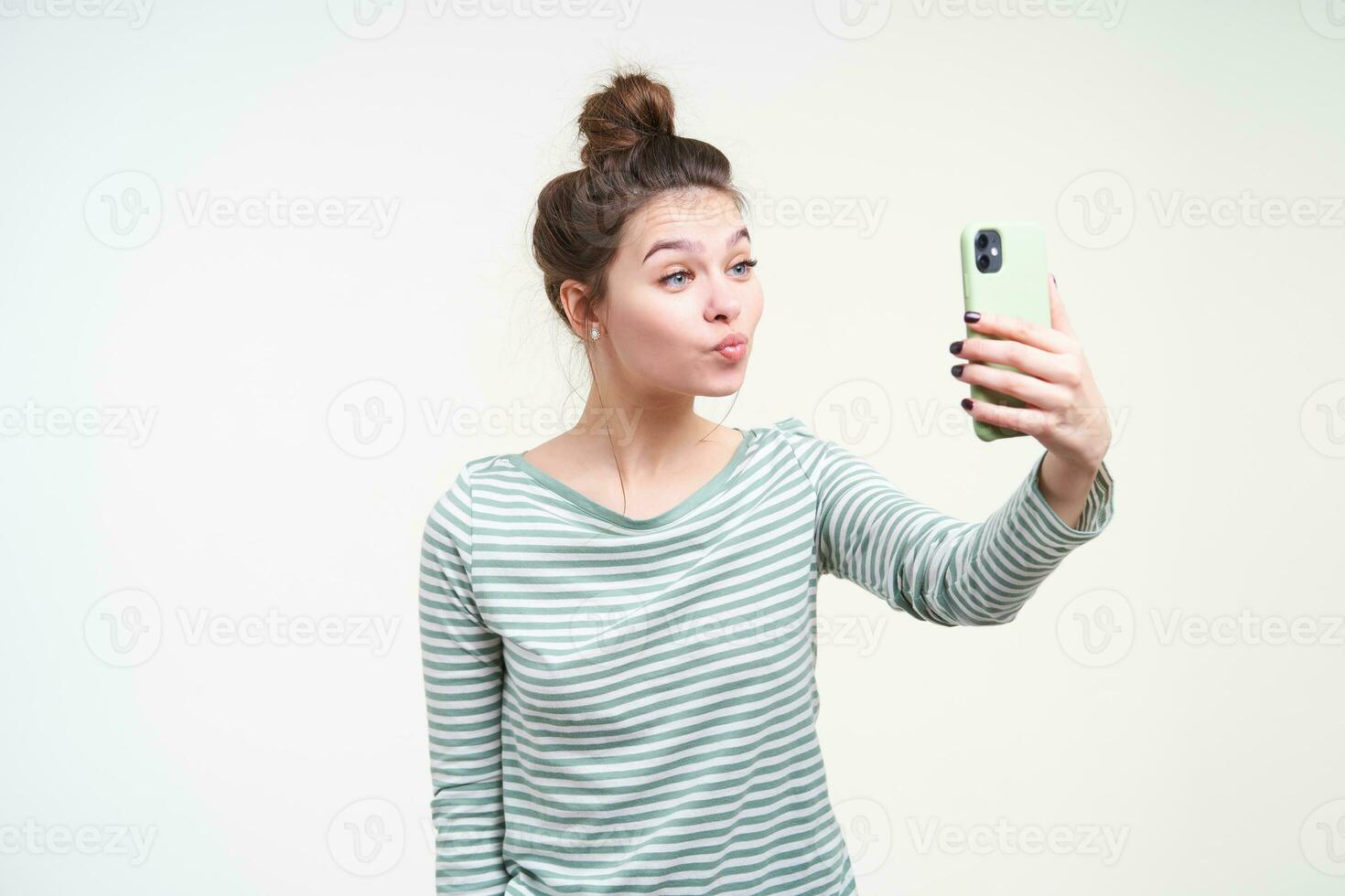 interno foto di giovane bello Marrone dai capelli signora con ciambella acconciatura imbronciato sua labbra mentre fabbricazione foto di se stessa e conservazione smartphone nel sollevato mano, isolato al di sopra di bianca sfondo