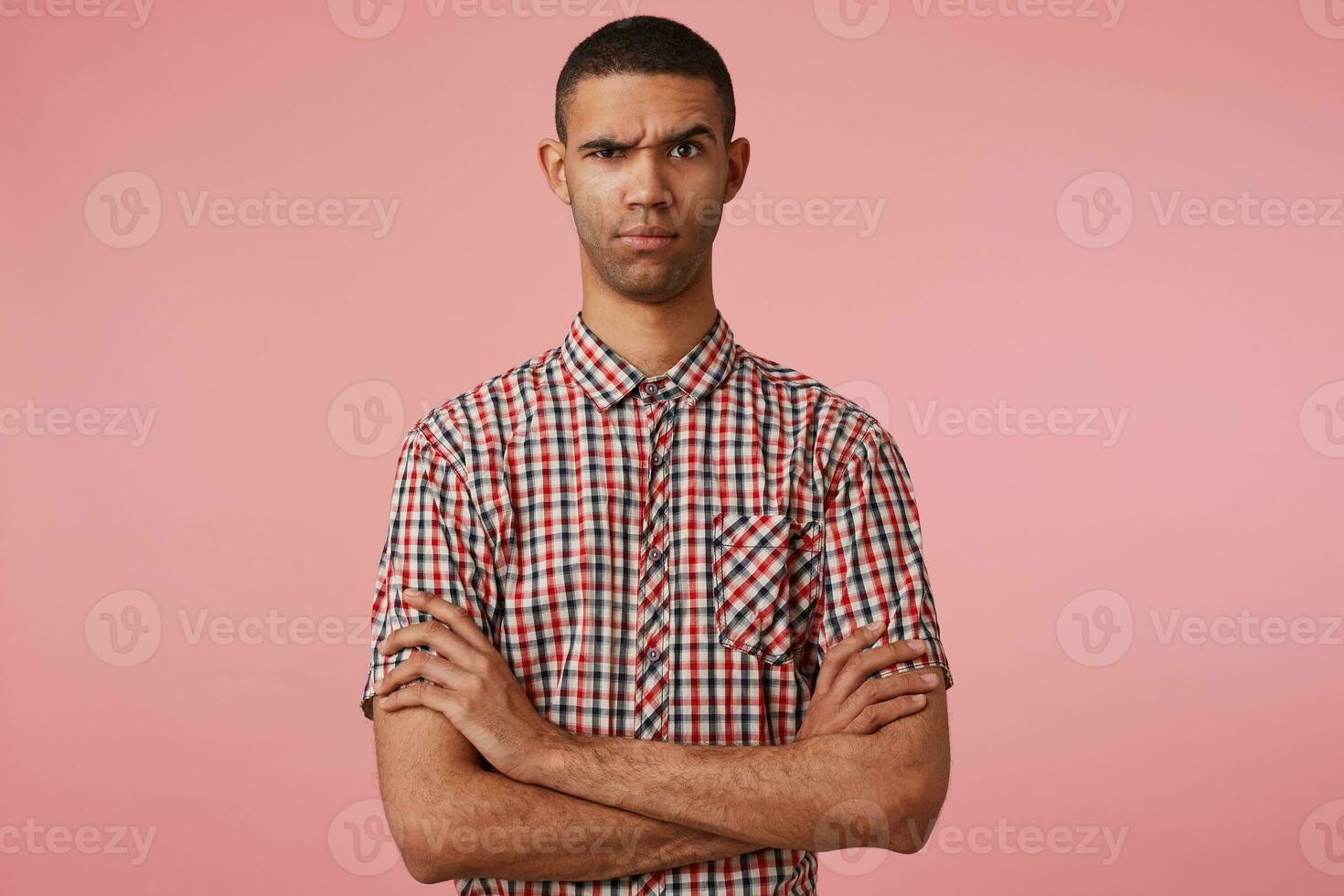 vicino su di giovane pdiscontentato buio spellato tipo nel scacchi camicia, malcontento sembra a il telecamera, sta al di sopra di rosa sfondo con incrociato braccia e sollevato sopracciglia. foto