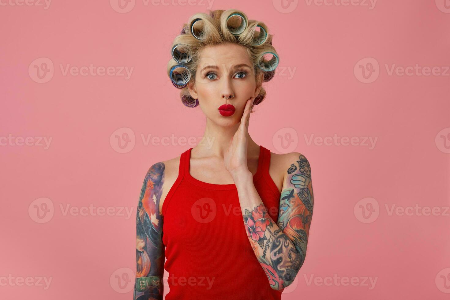 ritratto di attraente giovane bionda tatuato femmina con bigodini su sua testa indossare sera trucco mentre in posa al di sopra di rosa sfondo, conservazione mano su sua viso e pieghevole sua rosso labbra foto