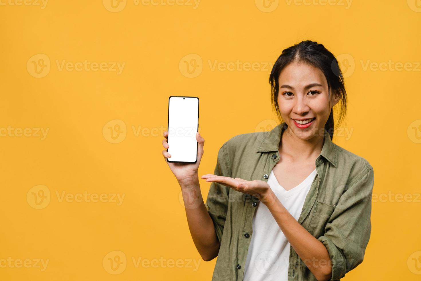 la giovane donna asiatica mostra lo schermo dello smartphone vuoto con un'espressione positiva, sorride ampiamente, vestita con abiti casual sentendosi felici su sfondo giallo. telefono cellulare con schermo bianco in mano femminile. foto