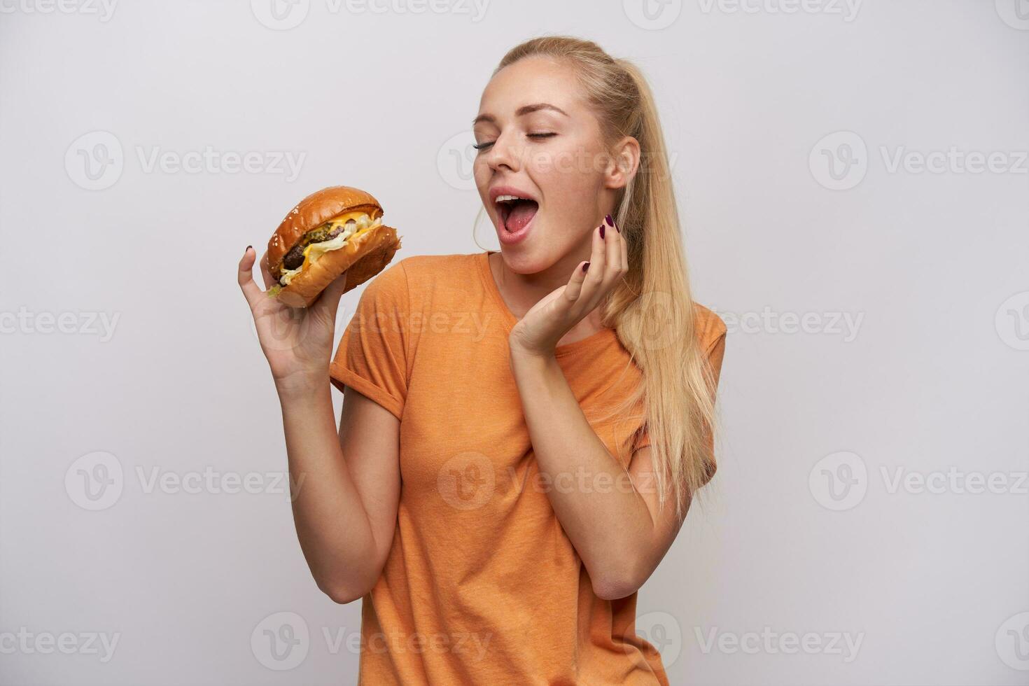 eccitato giovane bello lungo dai capelli bionda signora nel arancia maglietta pregustare delizioso cena mentre avendo Hamburger nel sollevato mano, sorridente piacevolmente mentre in posa al di sopra di bianca sfondo foto